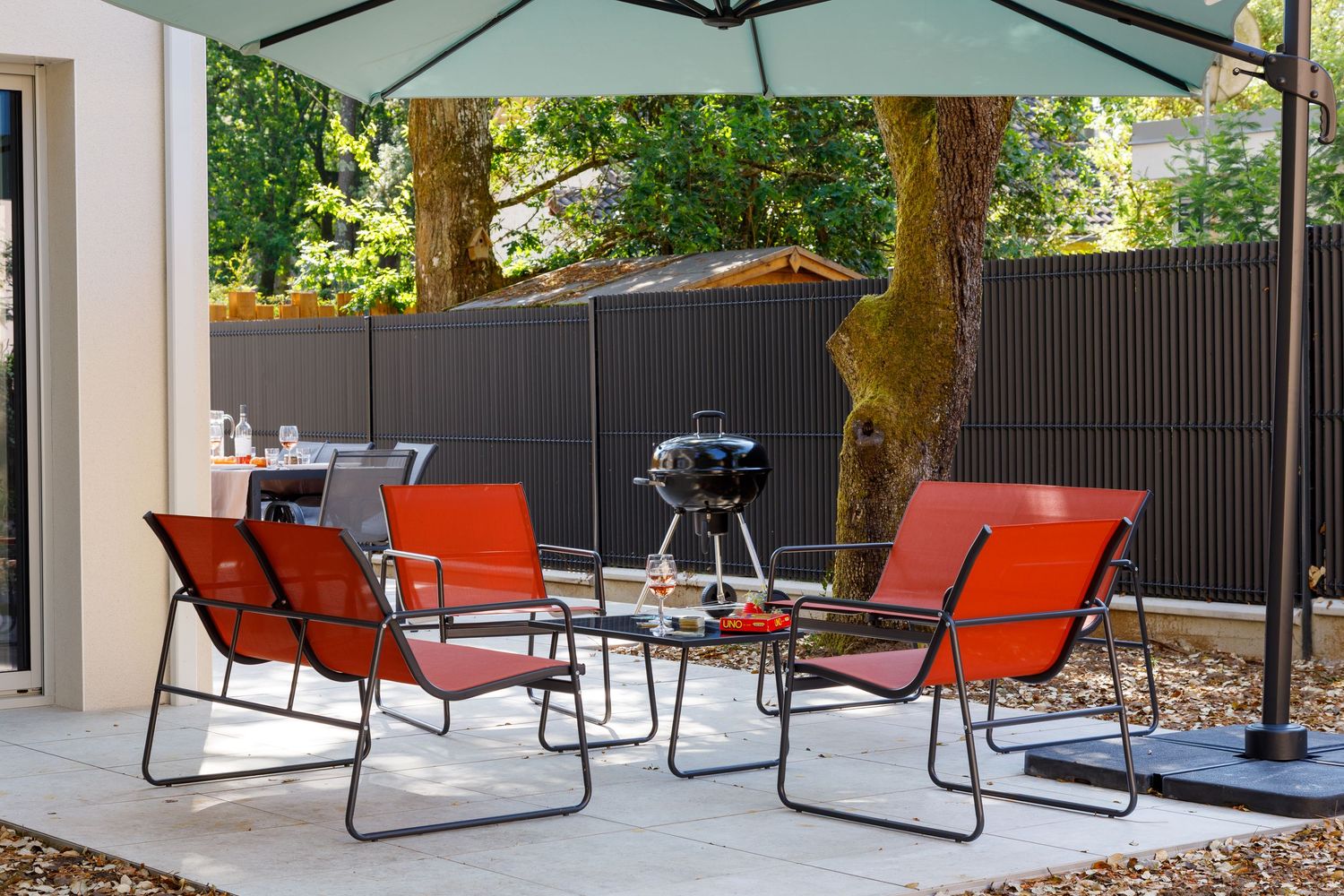 Terrasse extérieure avec des chaises rouges, une table en verre, un barbecue boule noir et un parasol, le tout sur une dalle en béton.