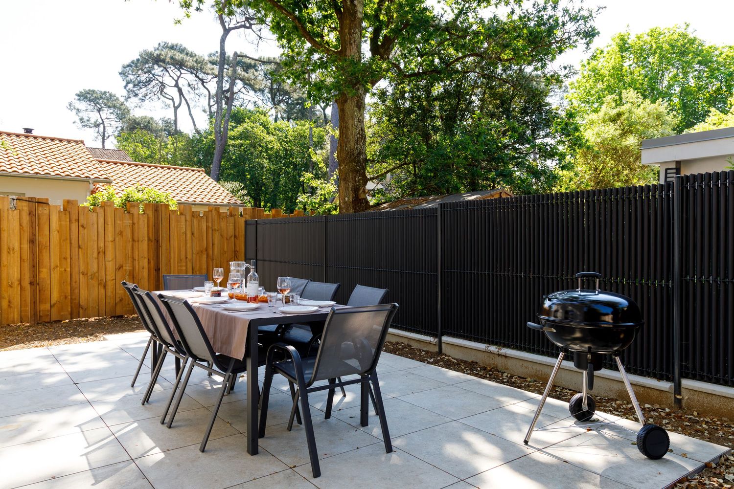 Terrasse extérieure avec table à manger, chaises et barbecue, entourée d'une clôture en bois et d'arbres verdoyants.