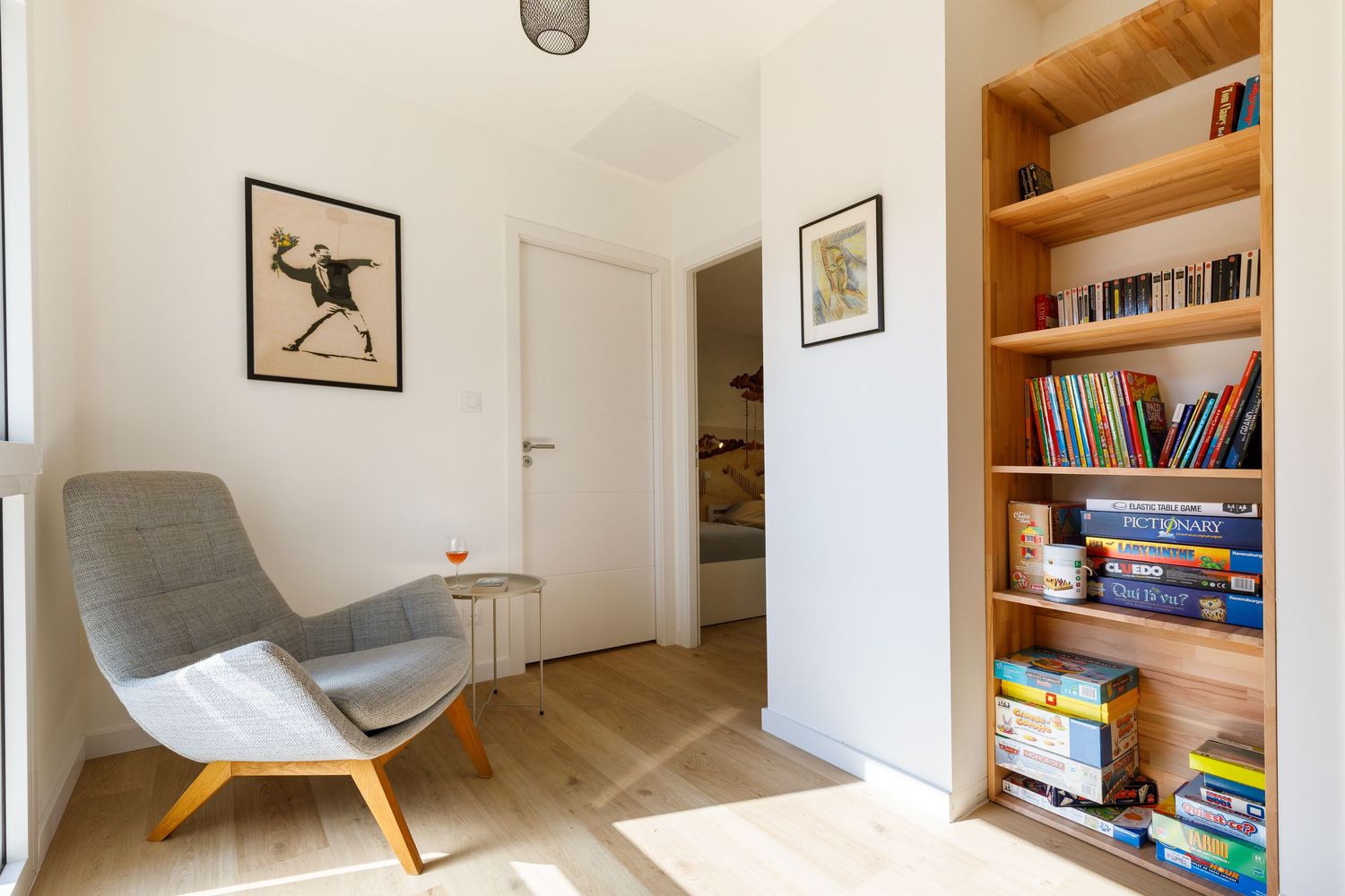 Coin lecture moderne avec un fauteuil gris, une reproduction d'œuvre de Banksy et une bibliothèque en bois intégrée remplie de livres coloré