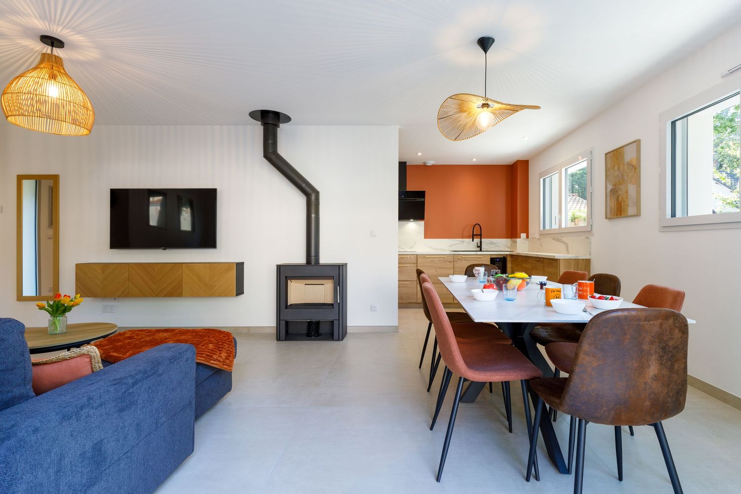 Séjour et salle à manger modernes avec poêle à bois, mur d'accent orange, suspensions en rotin et chaises en cuir brun.