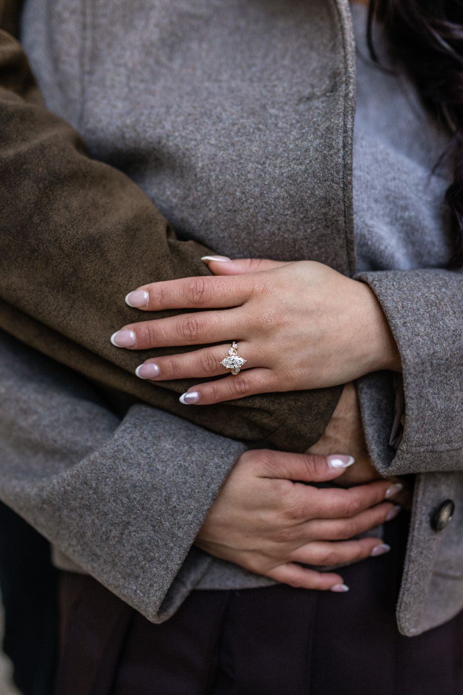 Close-up of a woman's hand with an engagement ring resting on a man's gray wool coat sleeve during an embrace.