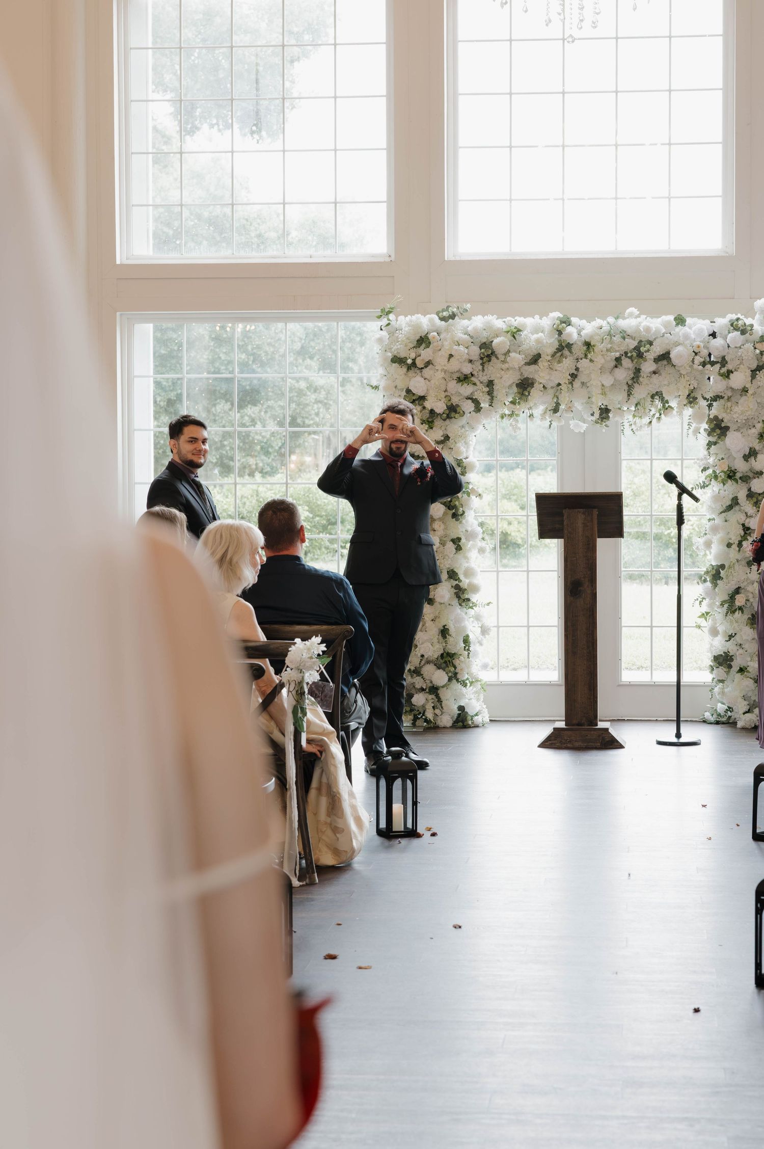 Officiant in a dark suit smiles at the end of an aisle beside a floral wedding arch and wooden podium in a bright, window-lined venue.