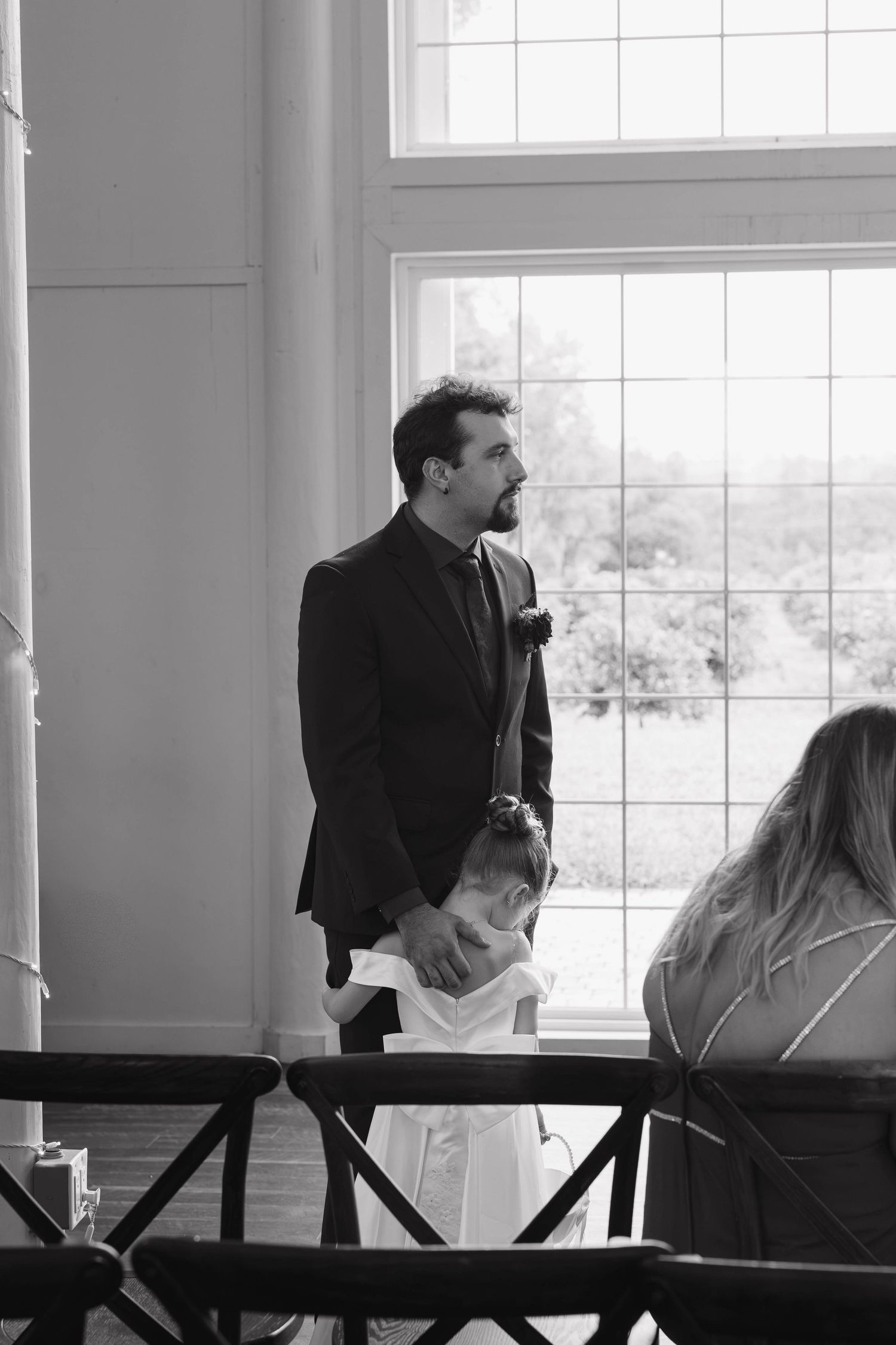 Black and white photo of a groom in a dark suit with a boutonniere standing near a large window, holding a flower girl's hand at a wedding.