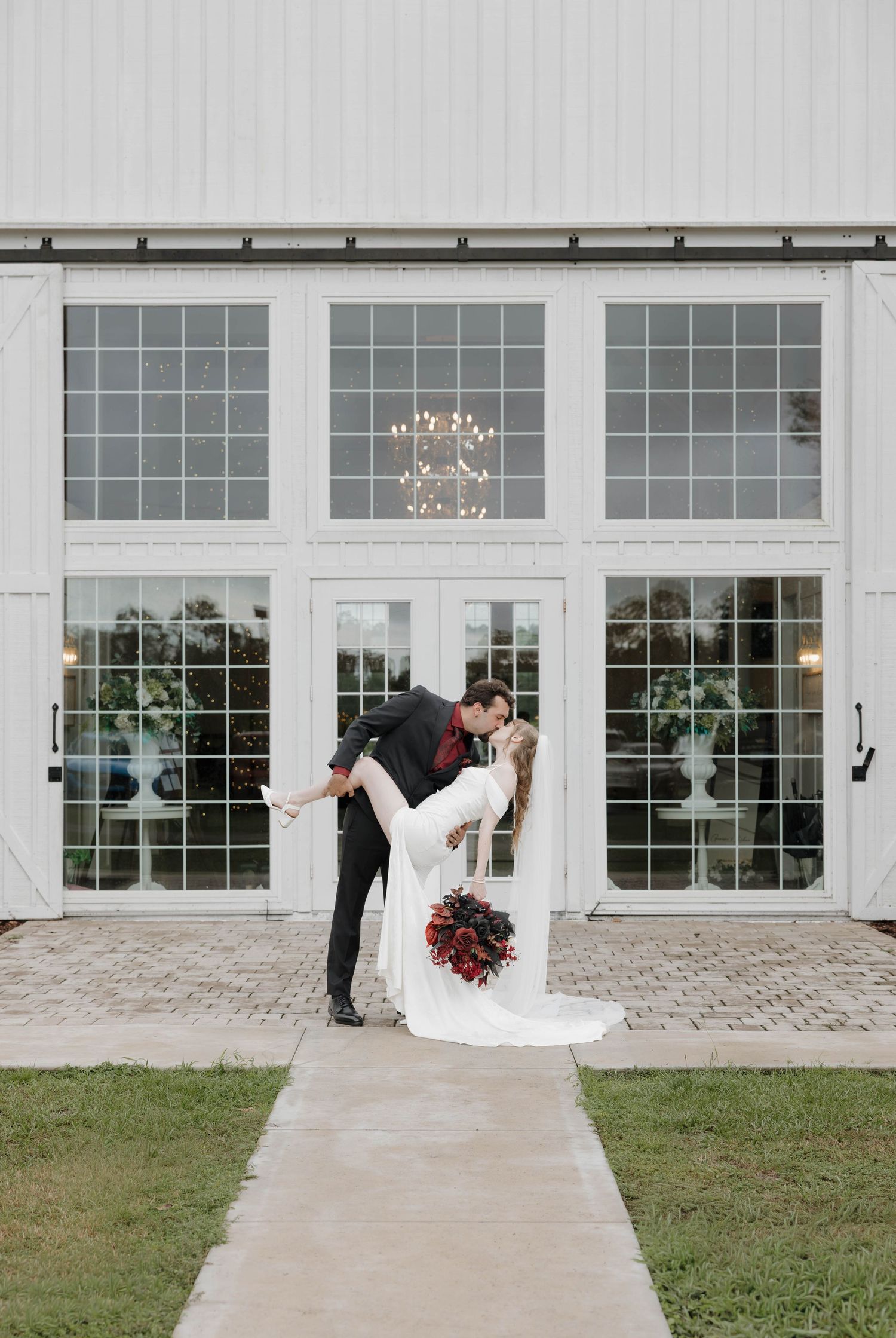 Groom dips and kisses his bride in a white gown holding a red bouquet outside a white barn wedding venue with large windows.