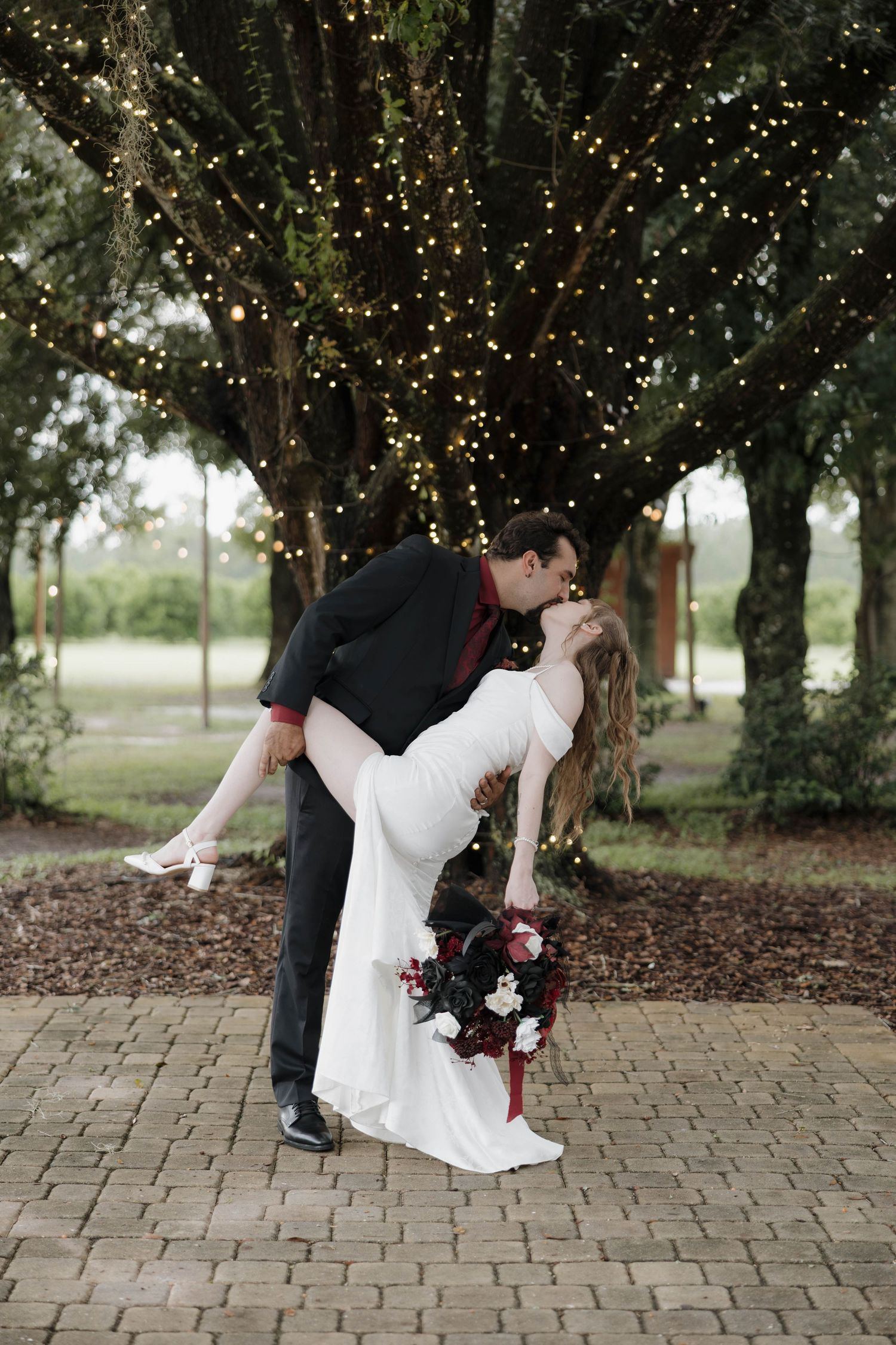 Groom dips and kisses bride in a white gown holding a dark red bouquet under oak trees draped in twinkling string lights.