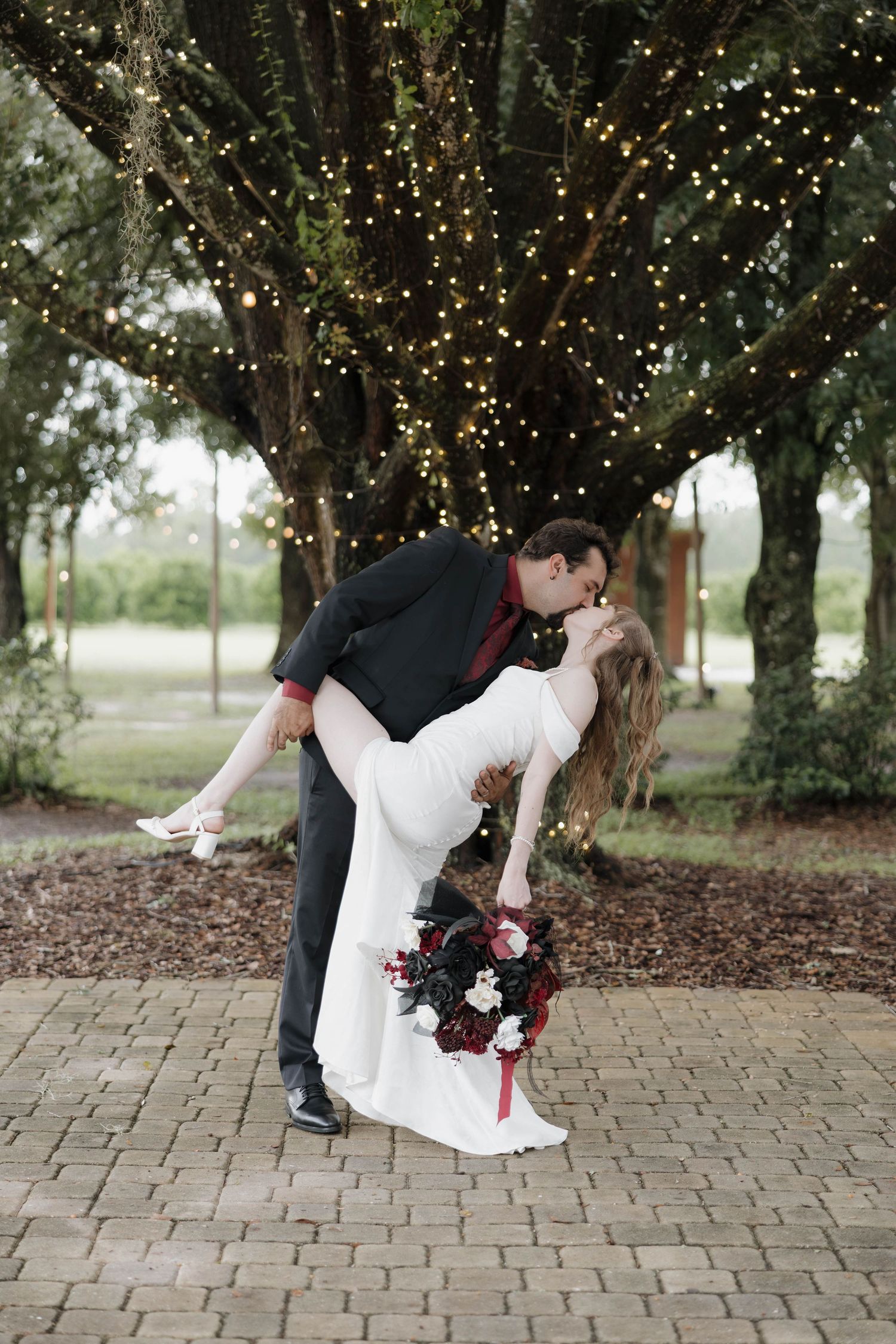 Groom dips and kisses bride in a white gown holding a red and white bouquet beneath an oak tree draped in string lights.