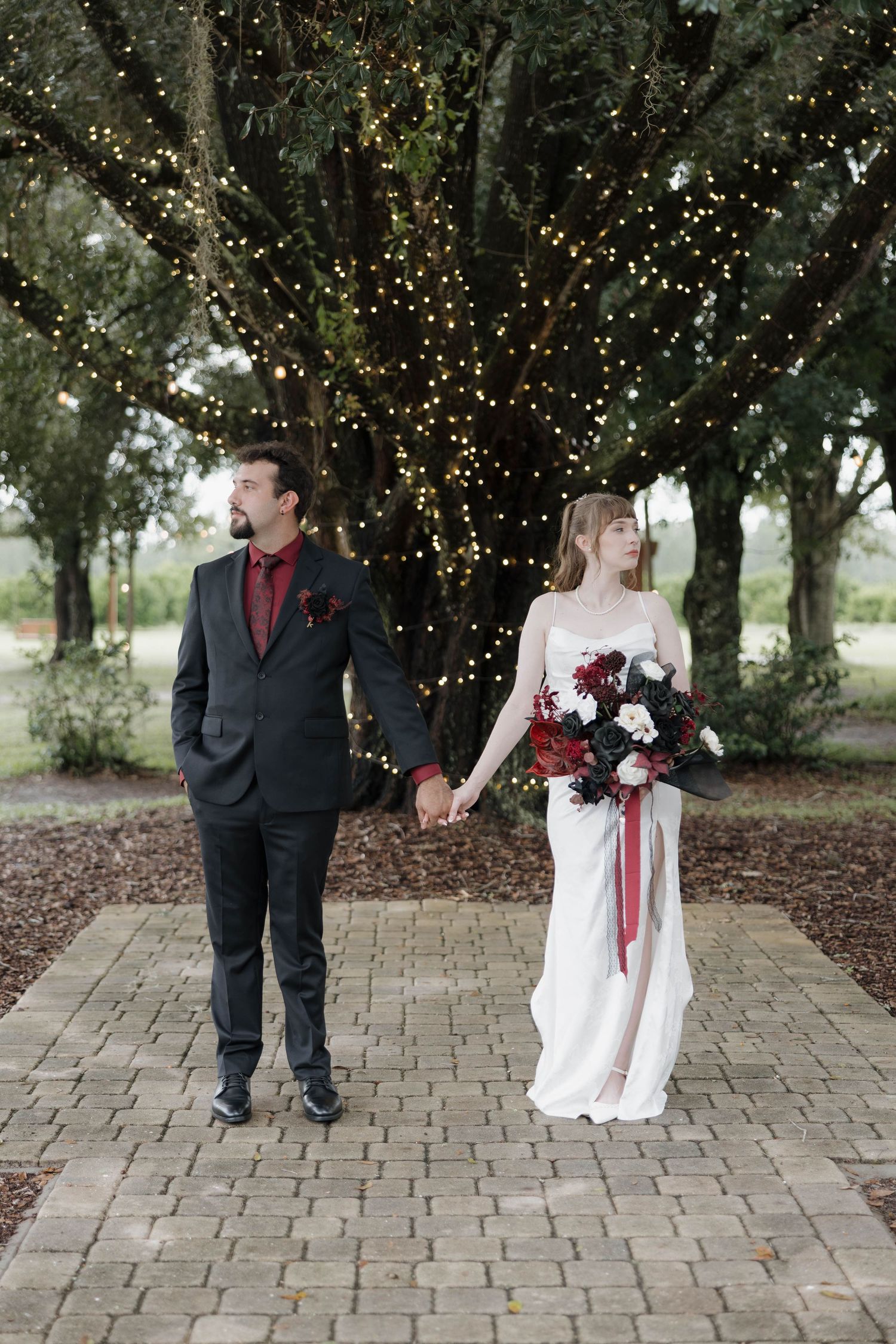 Bride and groom holding hands on a brick path beneath a string-lit tree, she holding a red and white bouquet at an outdoor wedding.