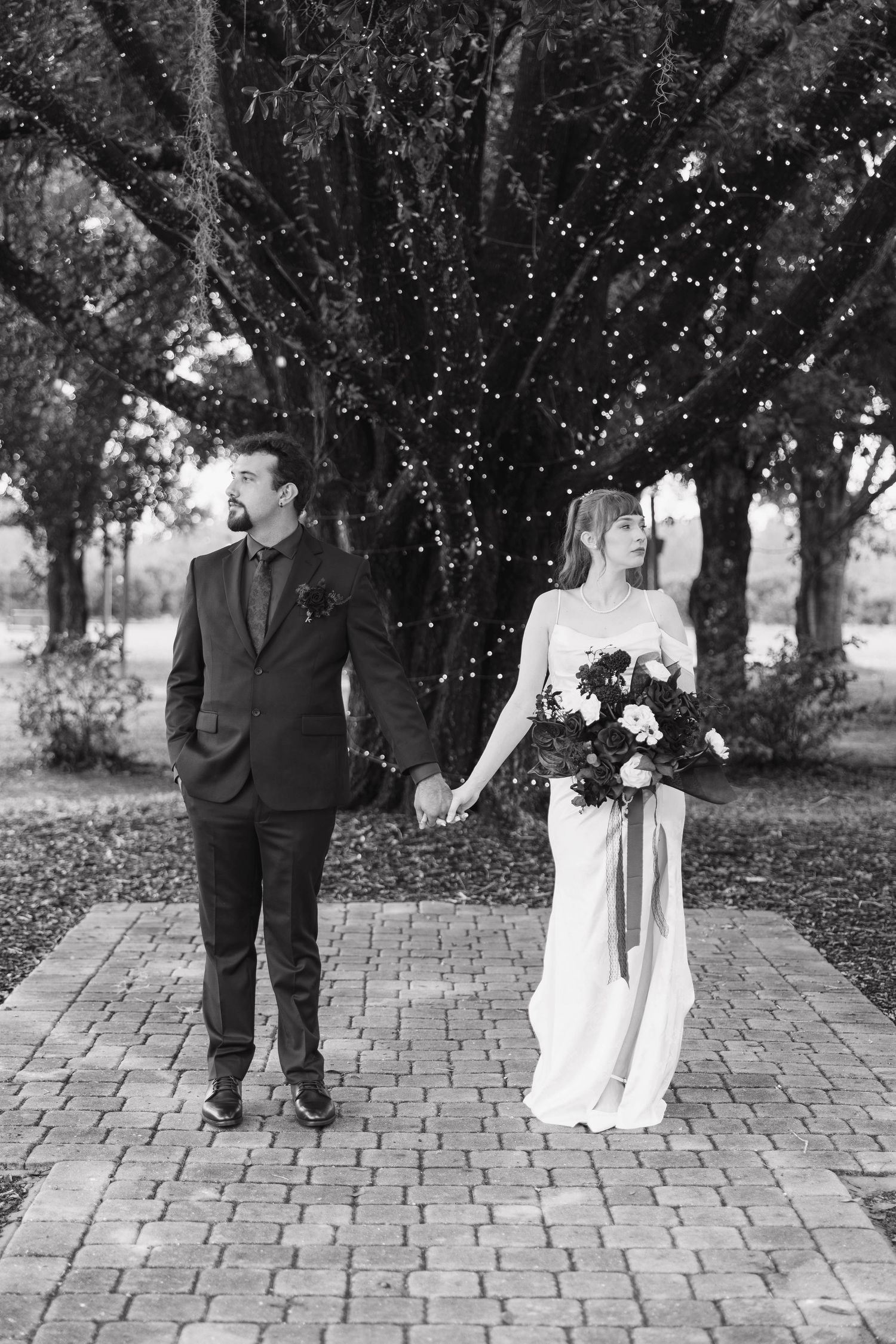 Bride and groom hold hands on a brick path beneath a large oak tree draped with string lights in a black and white wedding photo.