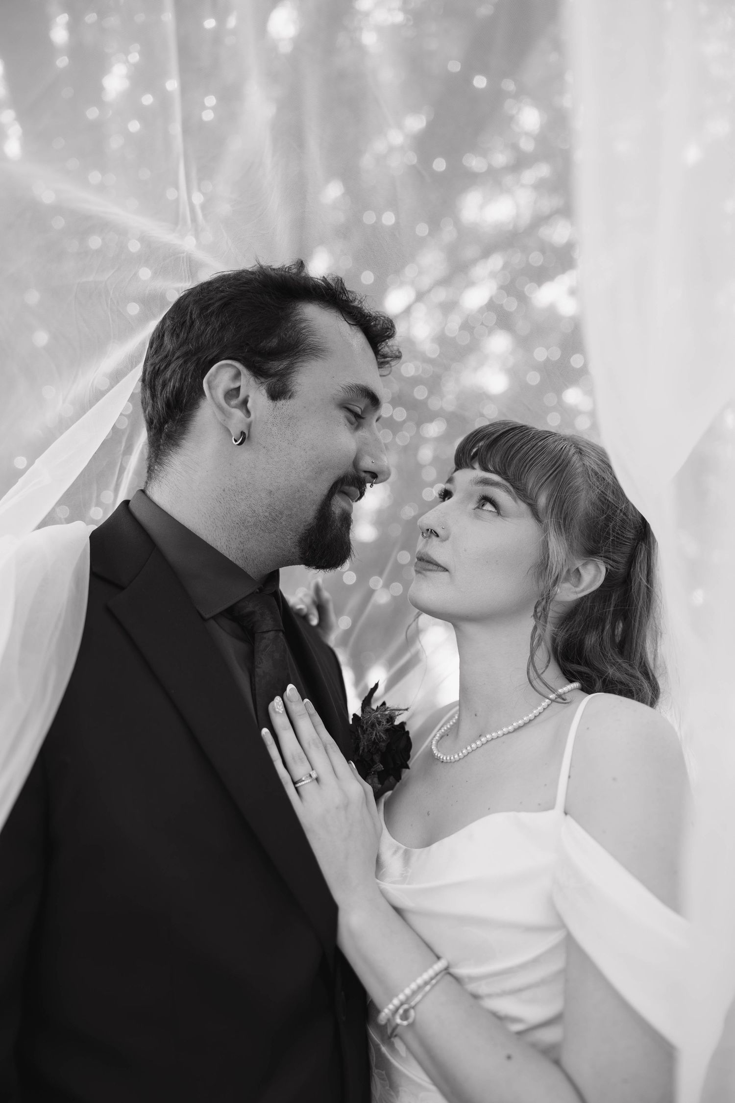 Black and white portrait of a bride and groom gazing at each other under a sheer veil, her hand resting on his lapel.