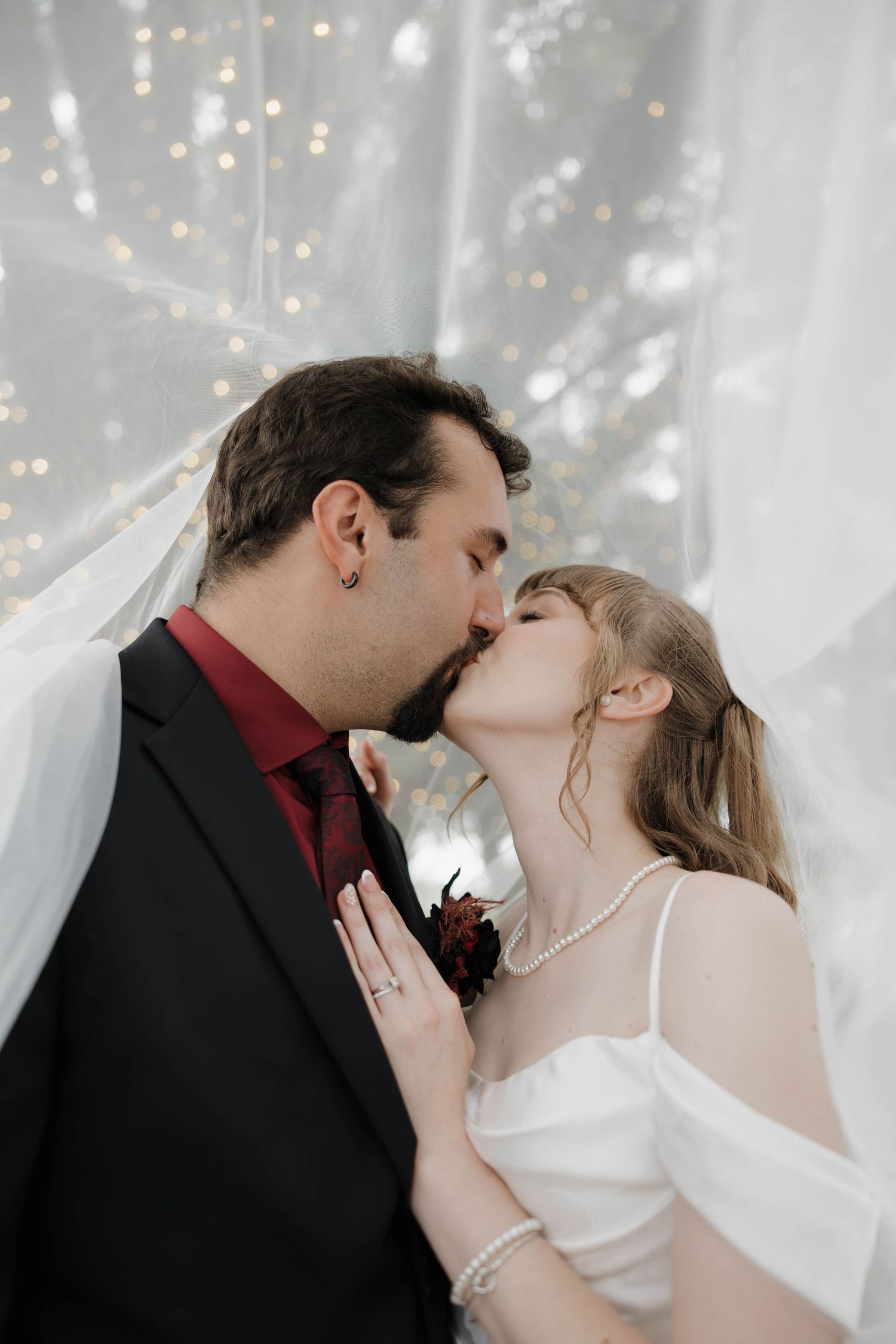 Bride and groom share a kiss under a sheer veil with twinkling string lights glowing in the background.