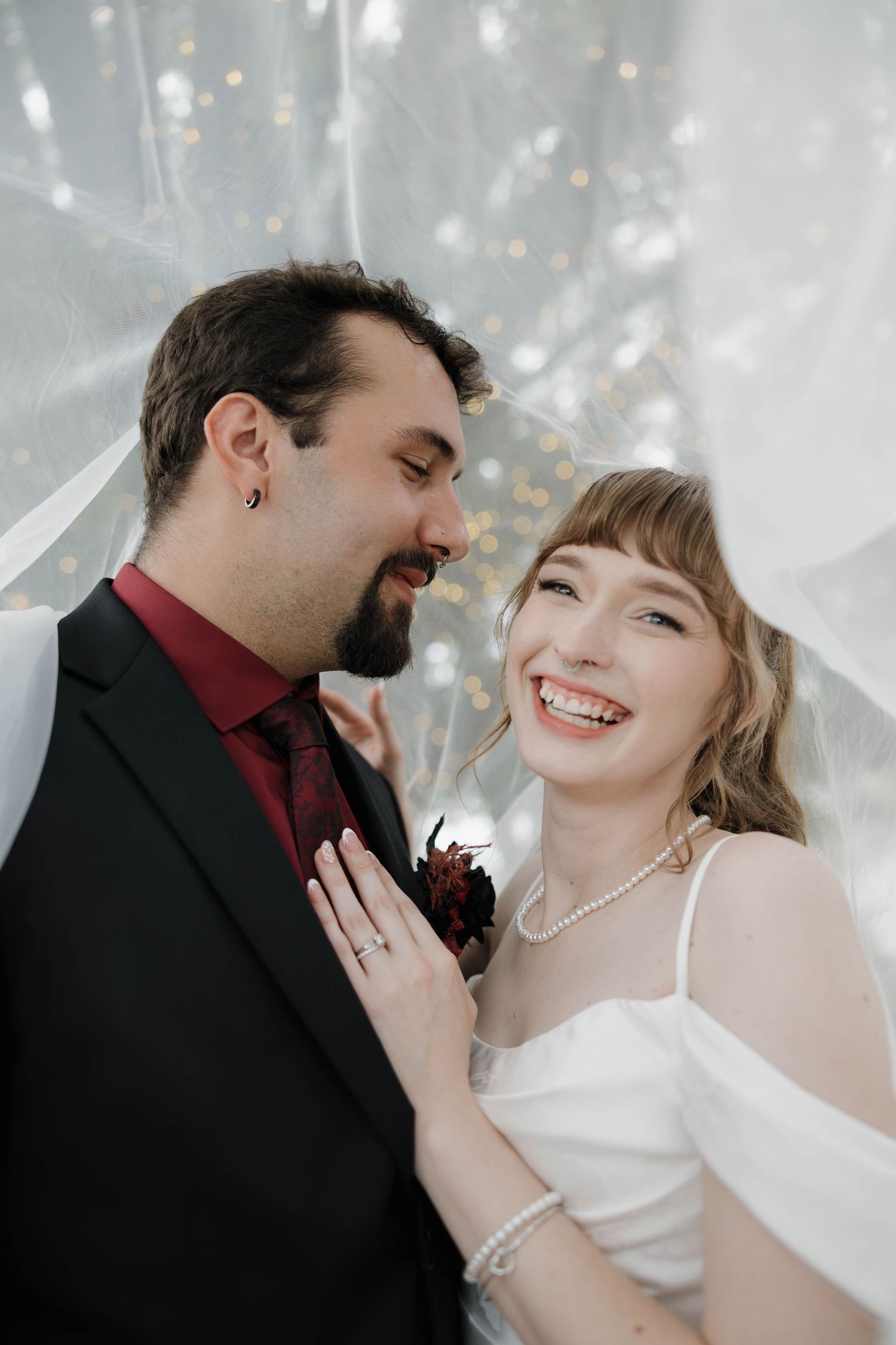 Smiling bride in a white satin dress and pearl necklace laughs beside a bearded groom in a black suit under a sheer veil.