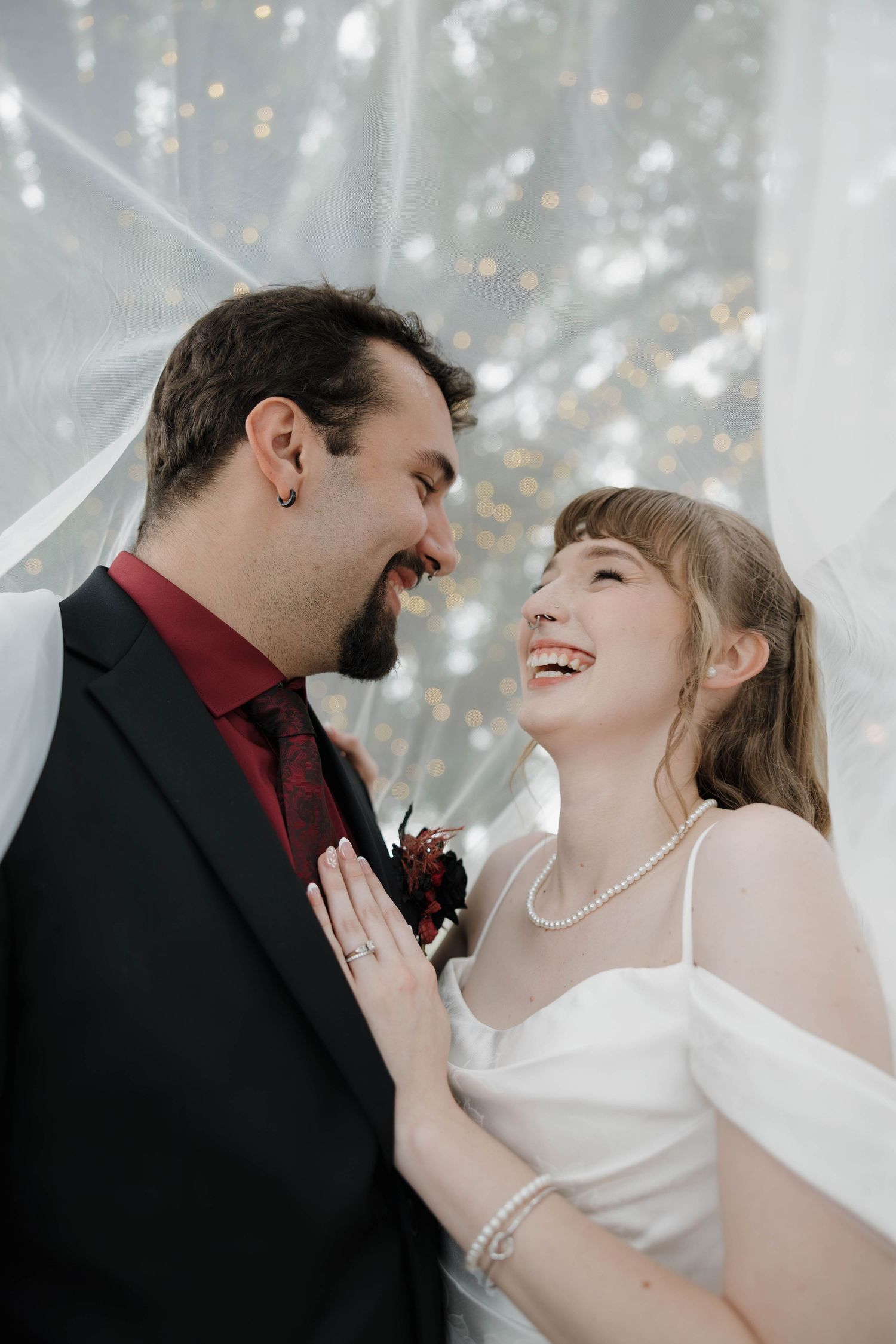 Laughing bride in a white gown and pearl necklace gazes at her smiling groom in a dark suit with a burgundy boutonniere under a sheer veil.