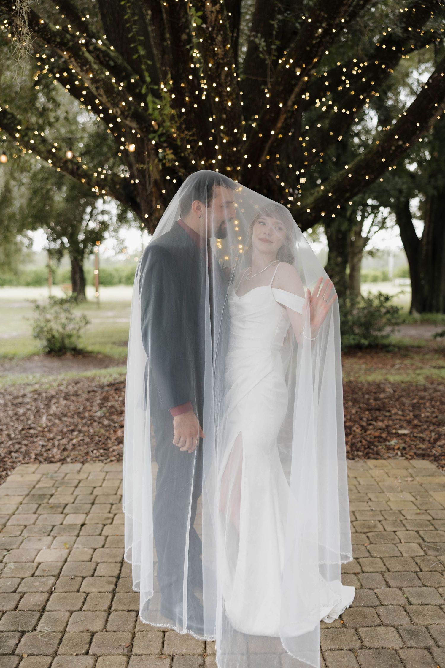 Bride and groom share a moment under a sheer wedding veil beneath a tree draped with twinkling string lights on a brick patio.