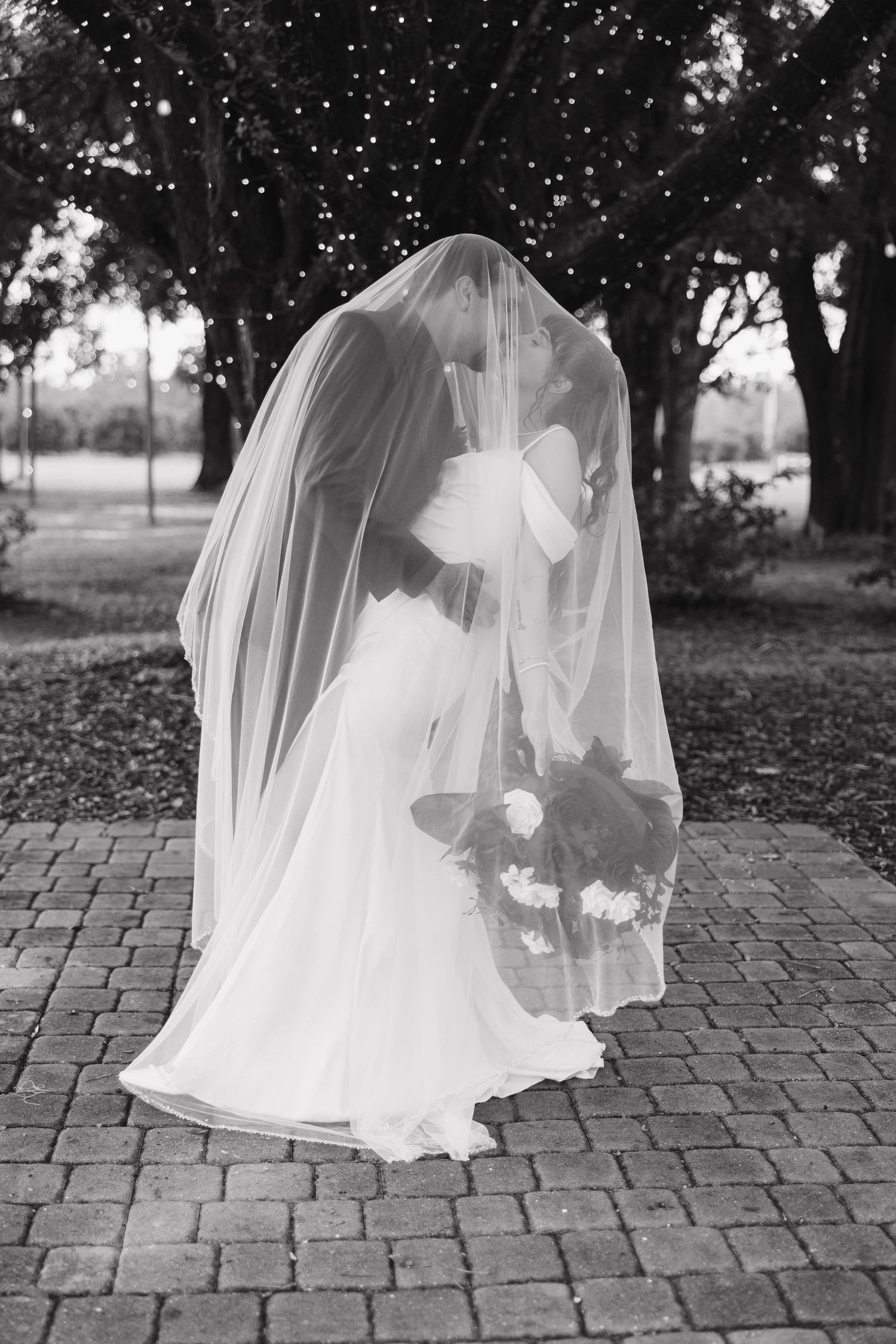 Black and white photo of a couple kissing beneath a long veil on a brick patio with string lights in the trees behind them.