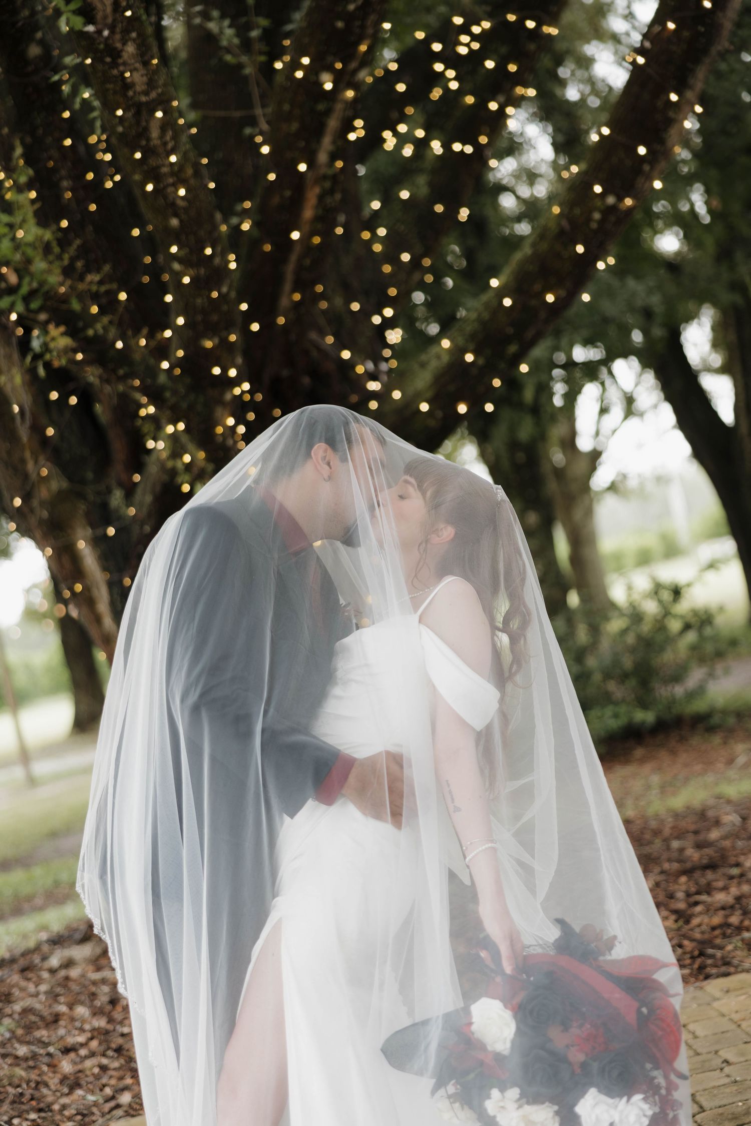 Bride and groom kiss beneath a sheer wedding veil holding a red and white bouquet under a tree draped with twinkling string lights.
