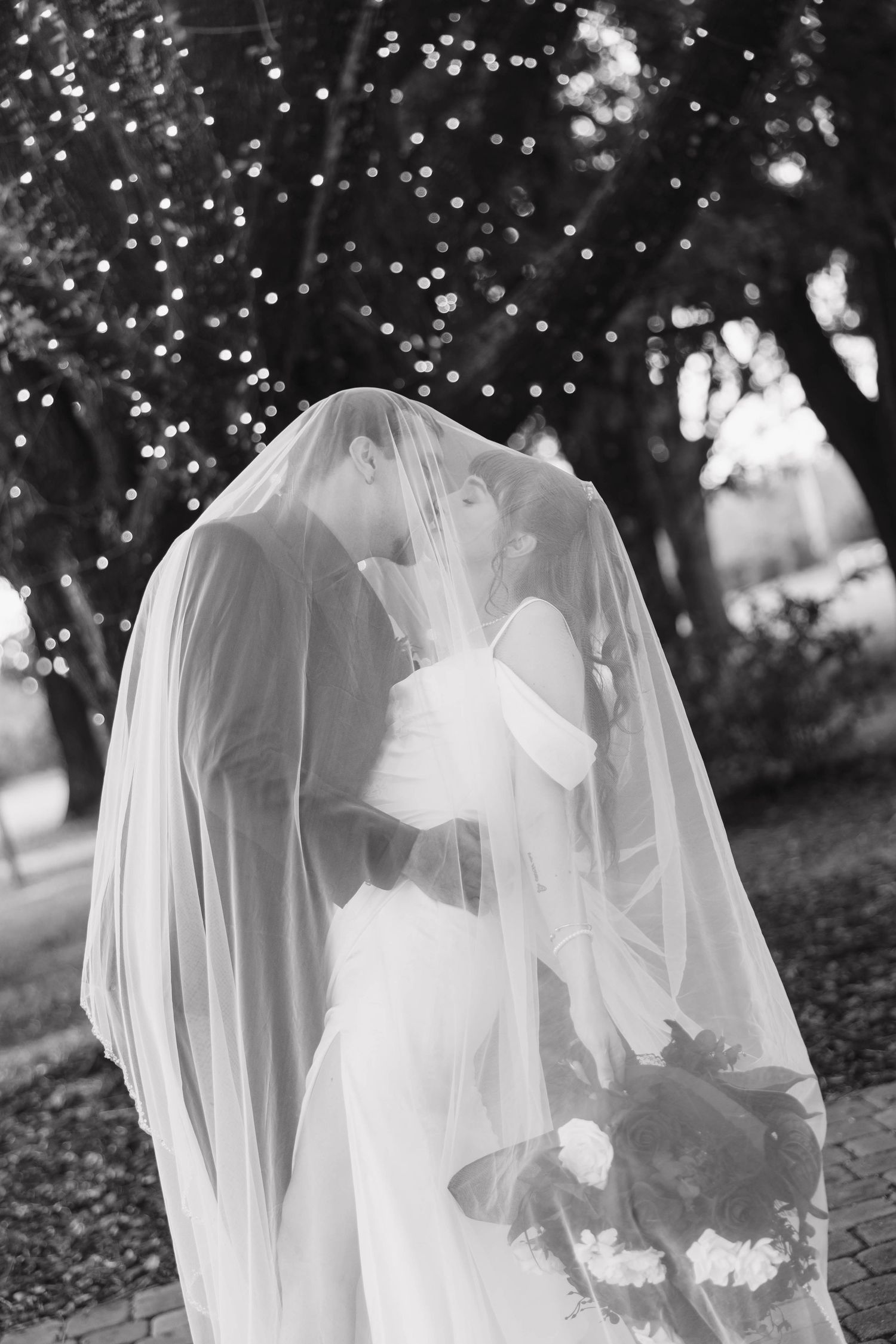 A black and white photo of a bride and groom kissing beneath a sheer veil, with sparkling string lights glowing in the trees behind them.