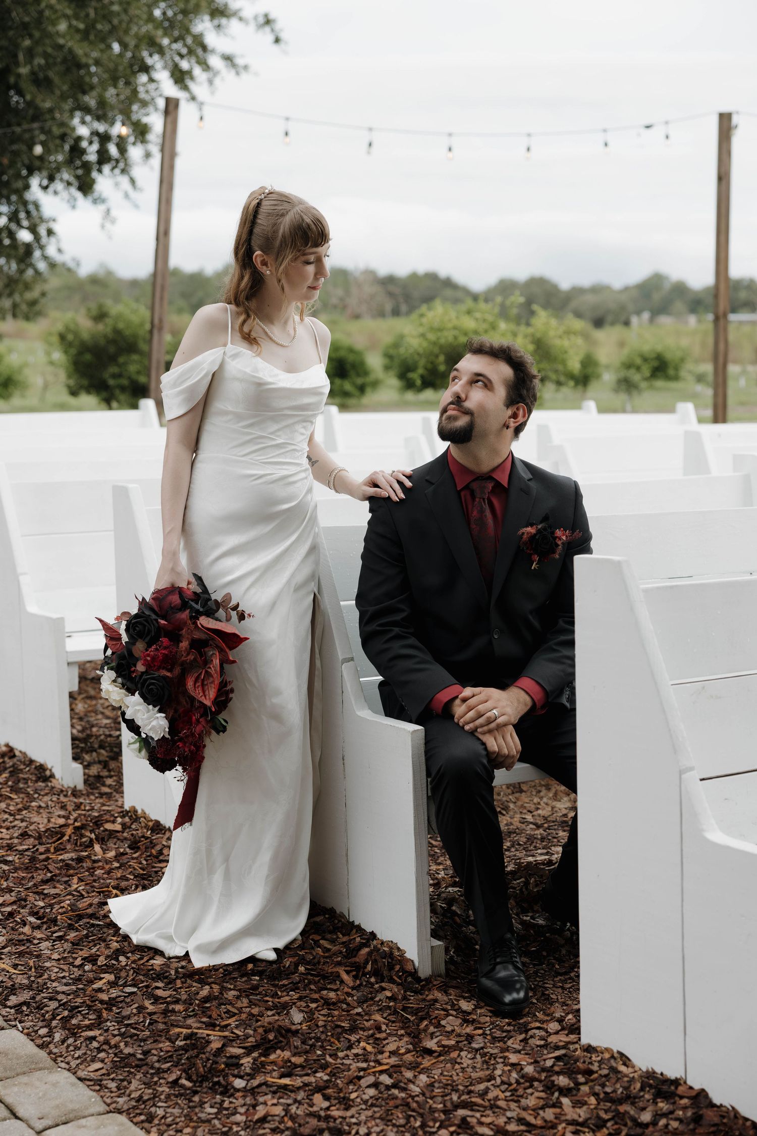 Bride in a white satin gown with a dark red bouquet rests her hand on a seated groom in a black suit at an outdoor wedding.