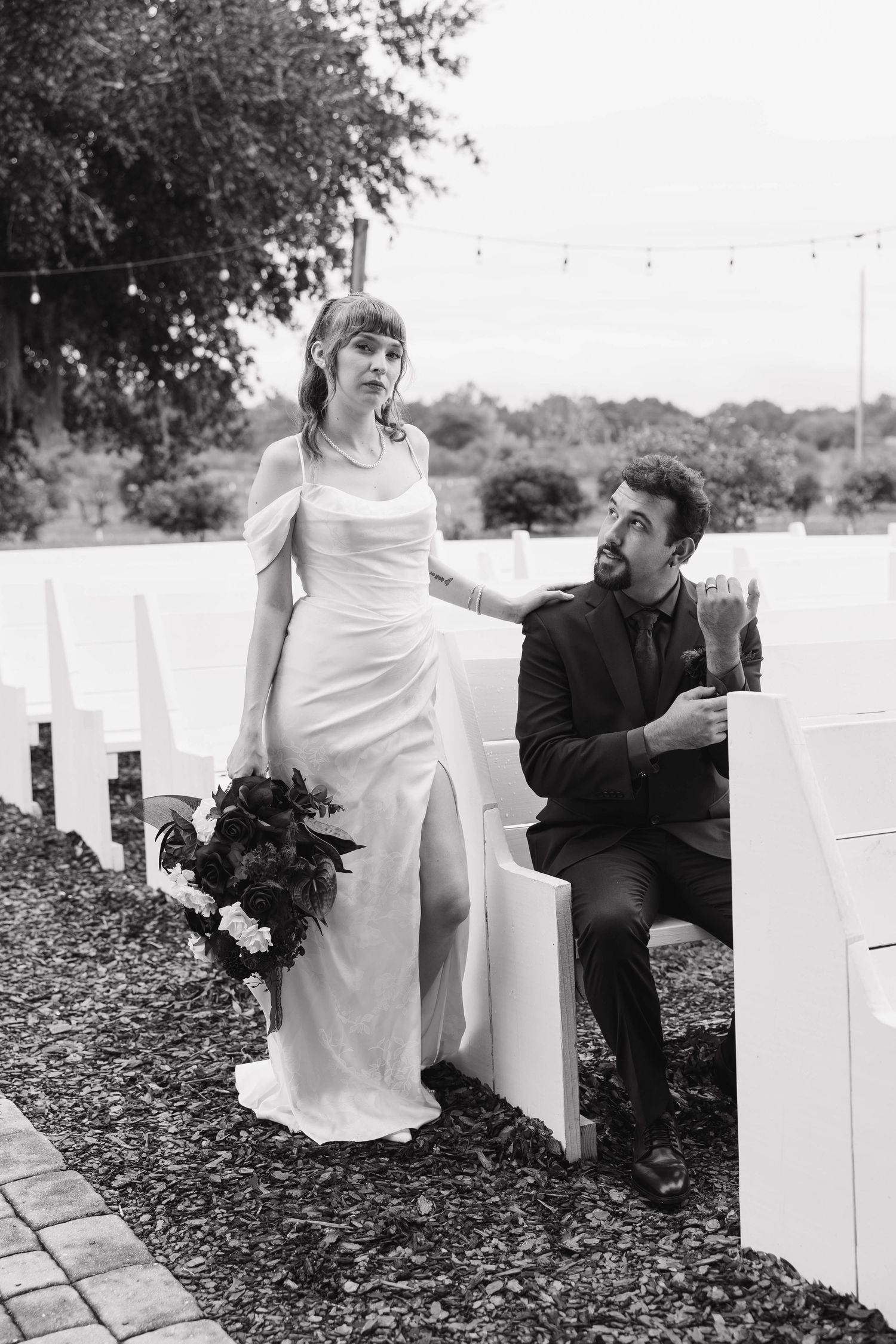 Black and white photo of a bride in a satin gown with a bouquet standing beside her groom seated on a white outdoor ceremony bench.