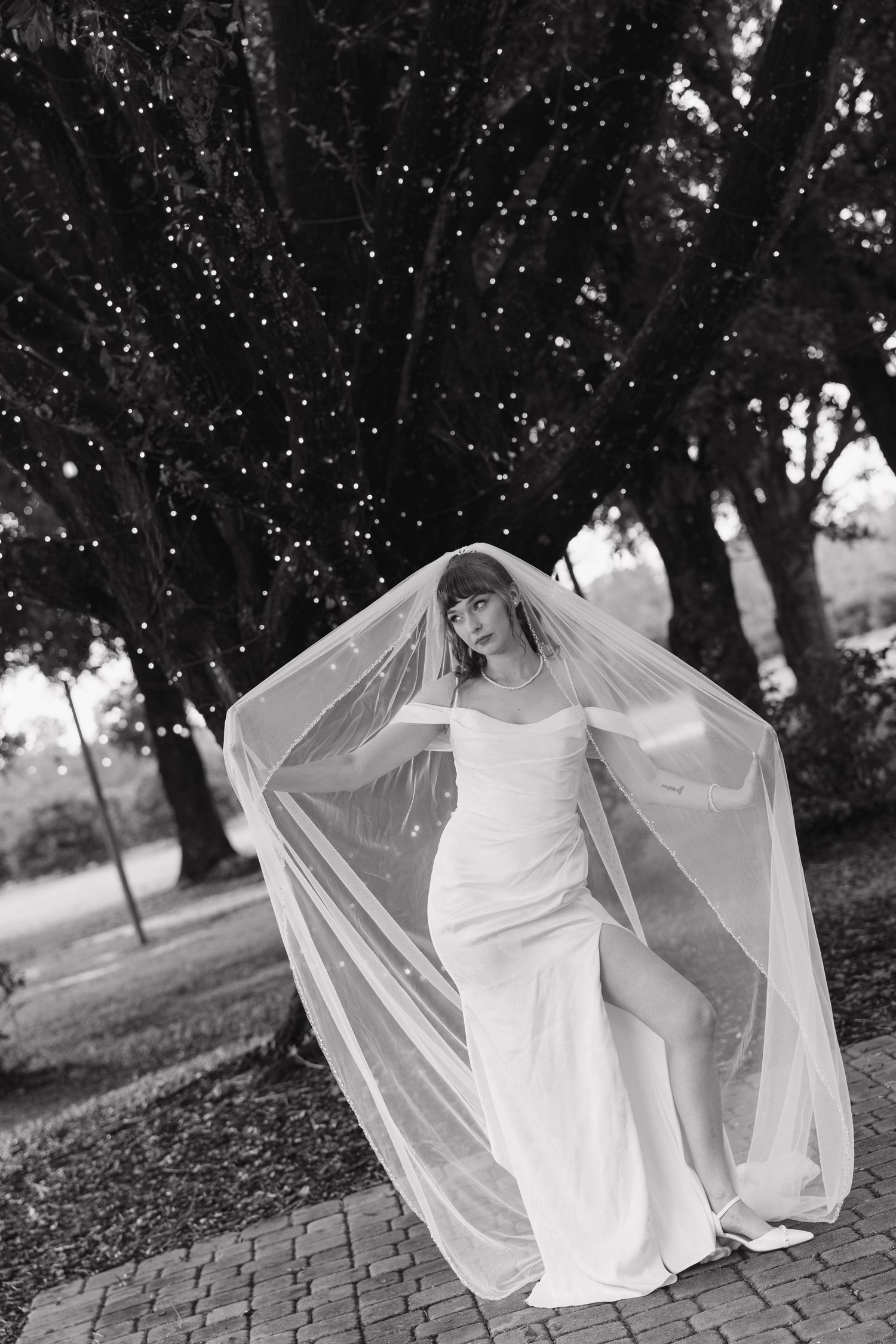 Black and white portrait of a bride in a fitted gown with a sheer veil standing under trees draped in string lights.