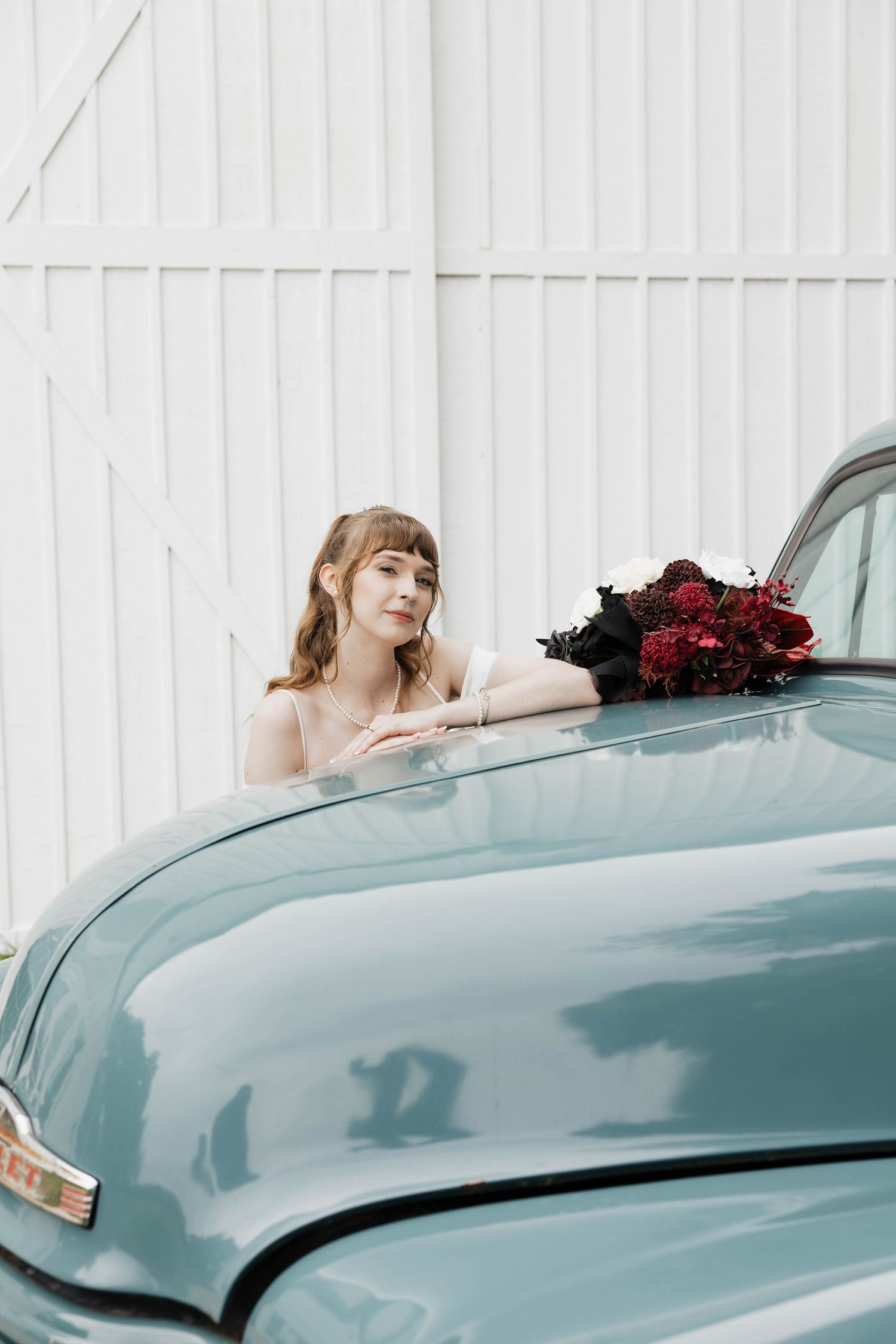 Bride in a white dress leans on the hood of a vintage teal truck beside a bouquet of deep red and white flowers.