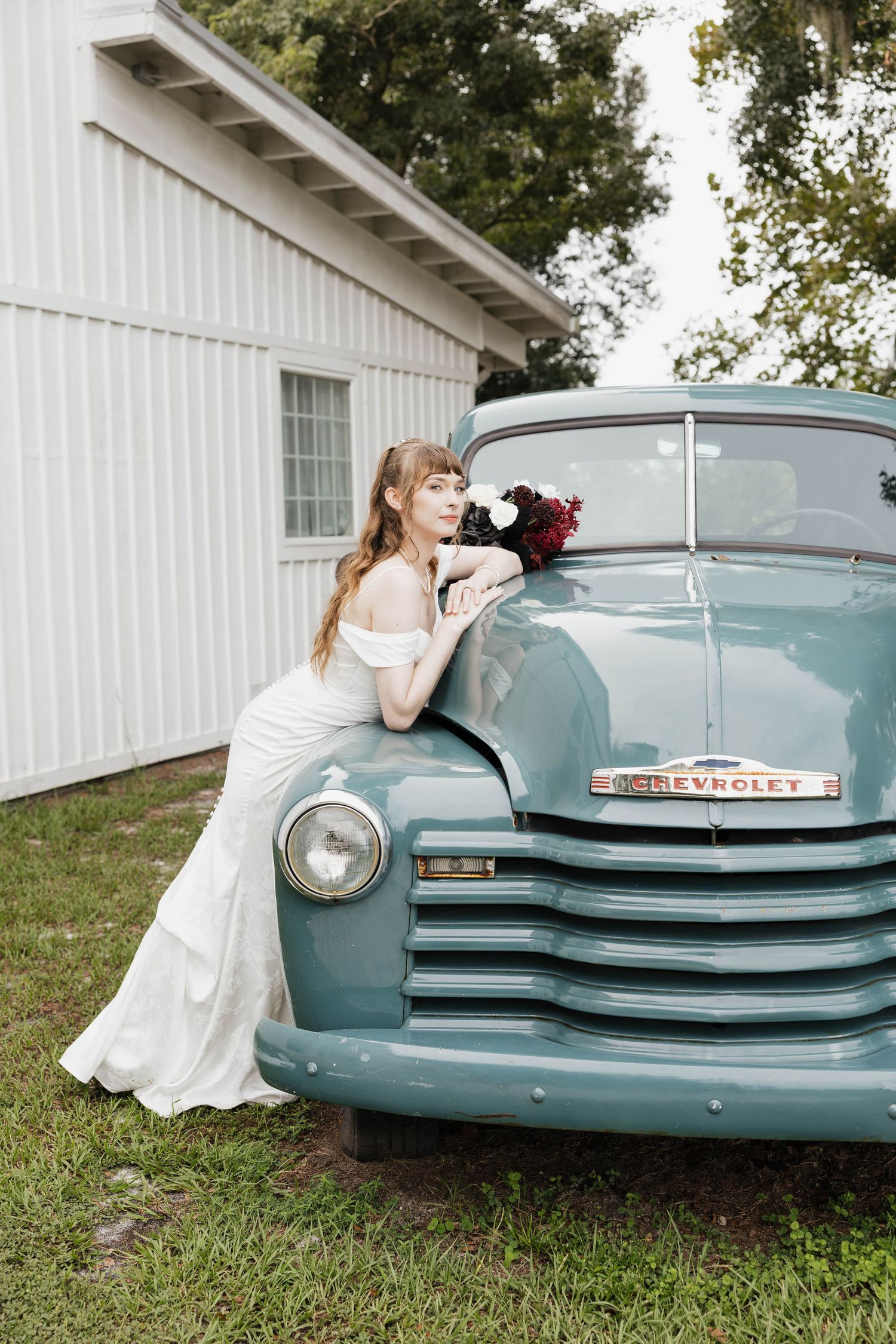 Bride in an off-the-shoulder white gown leaning on a vintage teal Chevrolet truck while holding a burgundy and white bouquet outdoors.