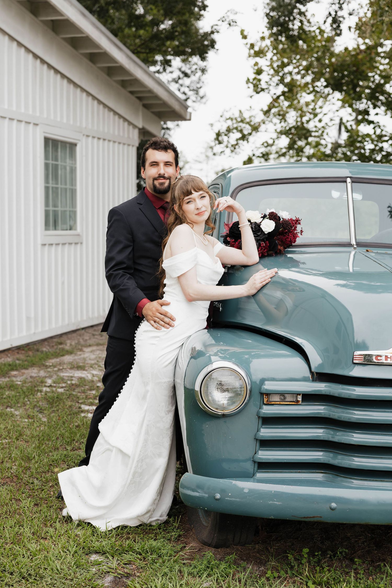 Bride in an off-shoulder white gown and groom in a dark suit pose beside a vintage teal pickup truck with a floral bouquet.