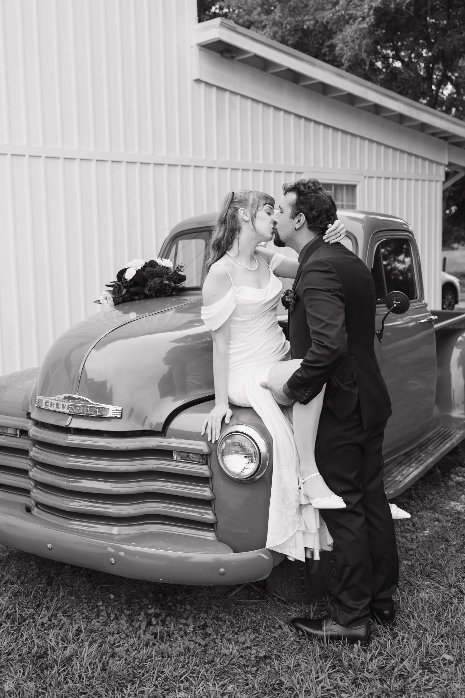 Black and white photo of a bride and groom kissing as she sits on the hood of a vintage Chevrolet truck at their wedding.