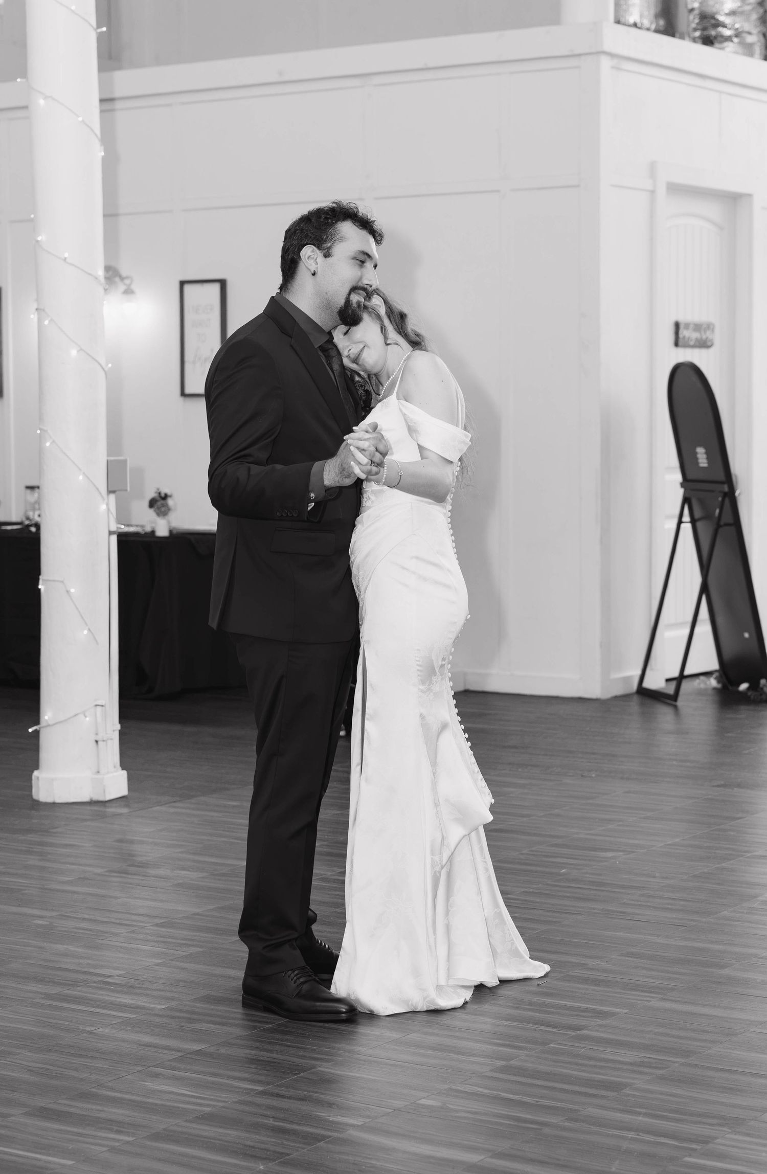 Black and white photo of the bride and groom sharing a slow first dance on a wood floor with string lights on a pillar.