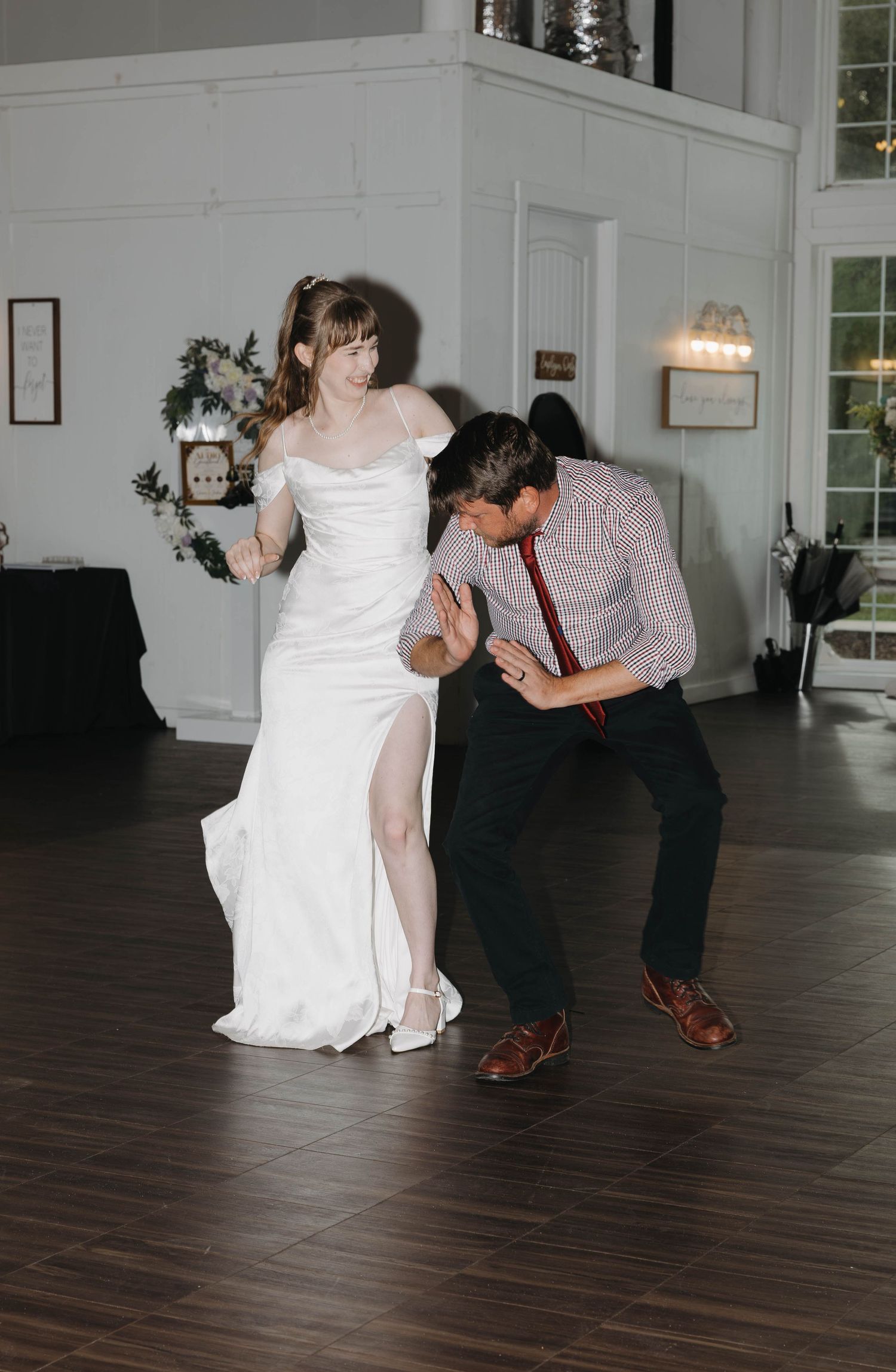 Bride in a white gown with a high slit laughs while dancing playfully with a guest in a plaid shirt and red tie on the dance floor.
