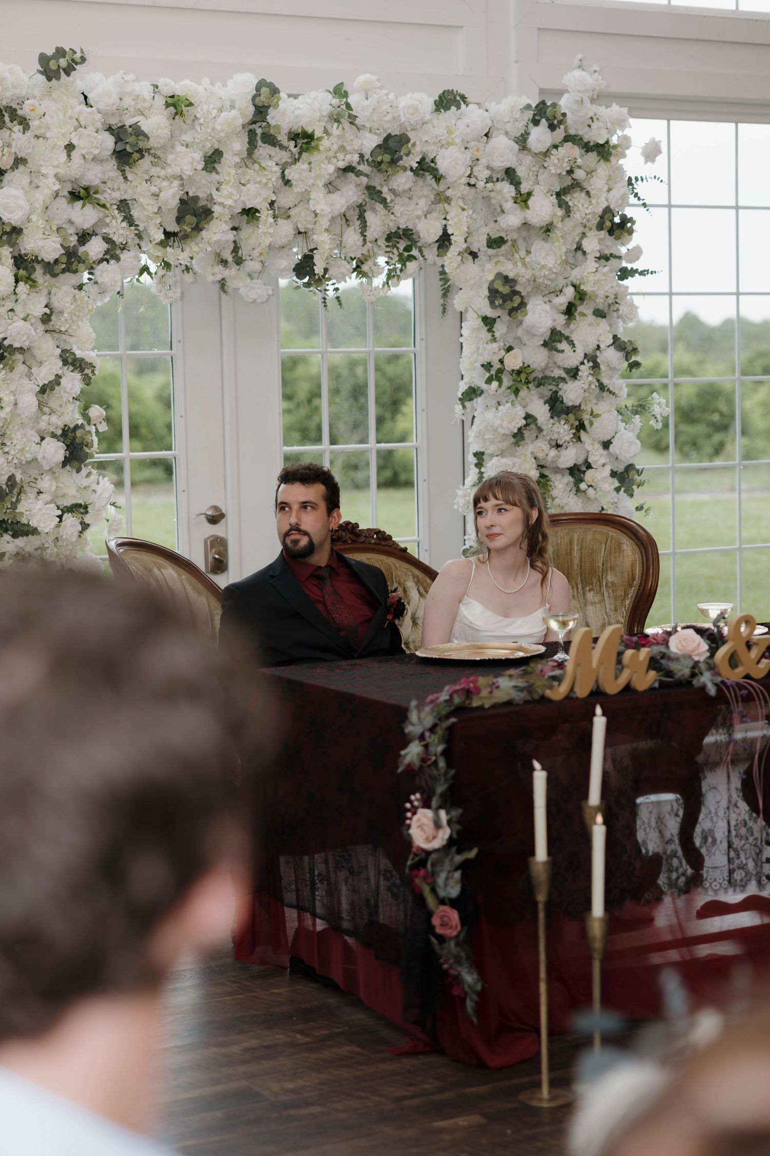Bride and groom sit at the sweetheart table beneath a lush white floral arch with gold letters and candles in front.