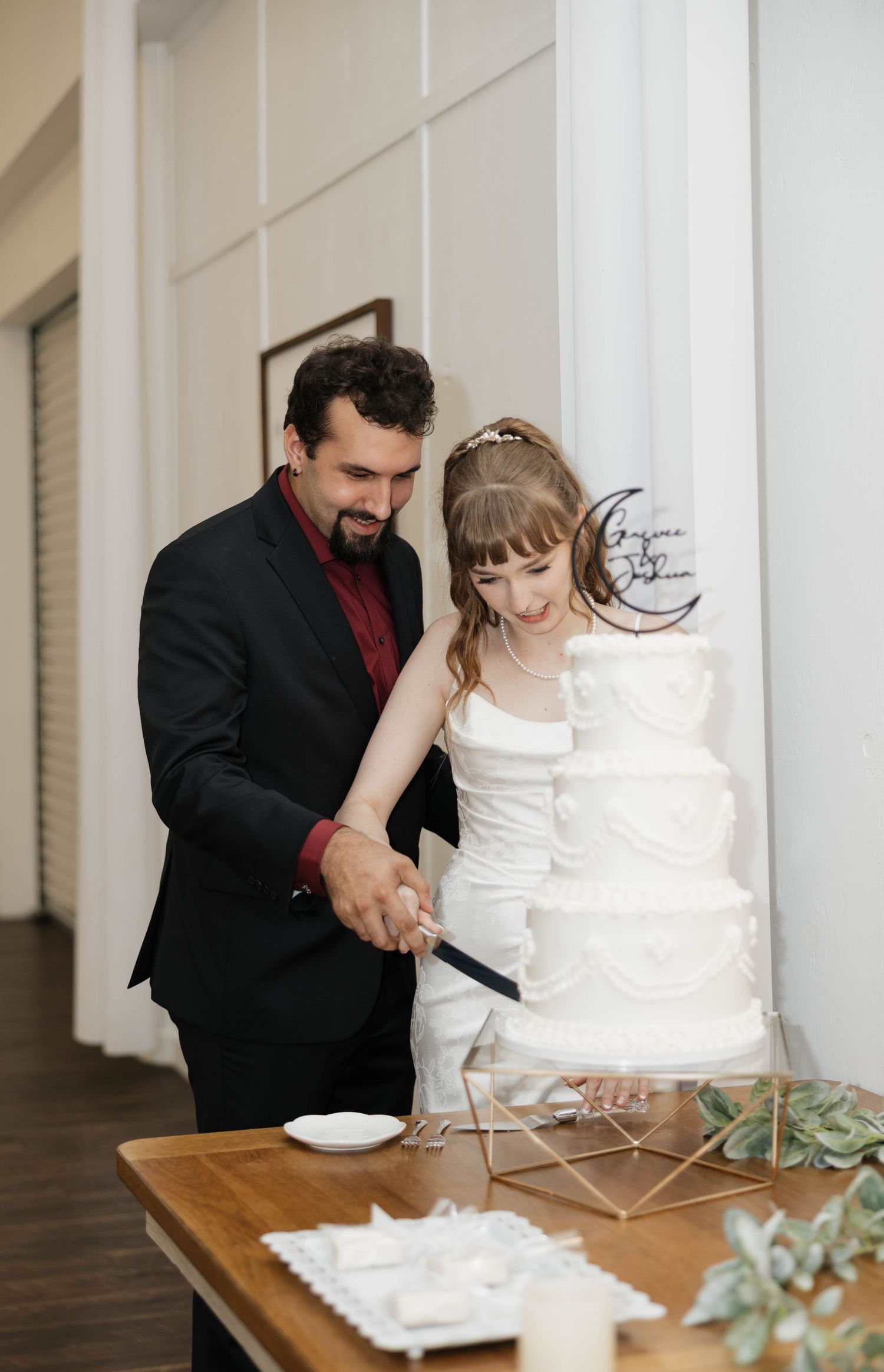 A bride in a white dress and a groom in a black suit cut their white tiered wedding cake together at a wooden table.