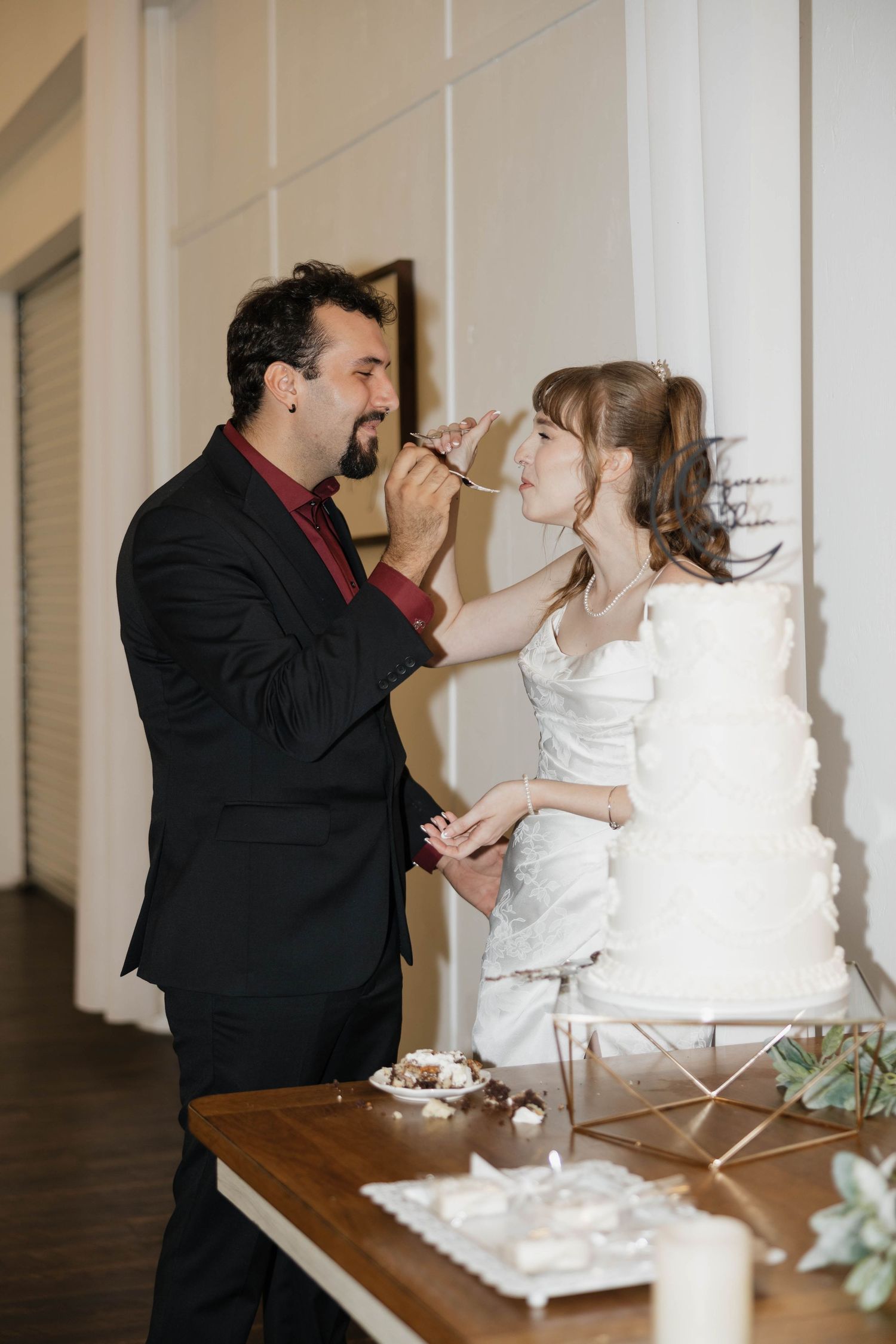 Groom in a black suit feeds a bite of cake to the bride in a white gown beside a tiered white wedding cake on a wooden table.