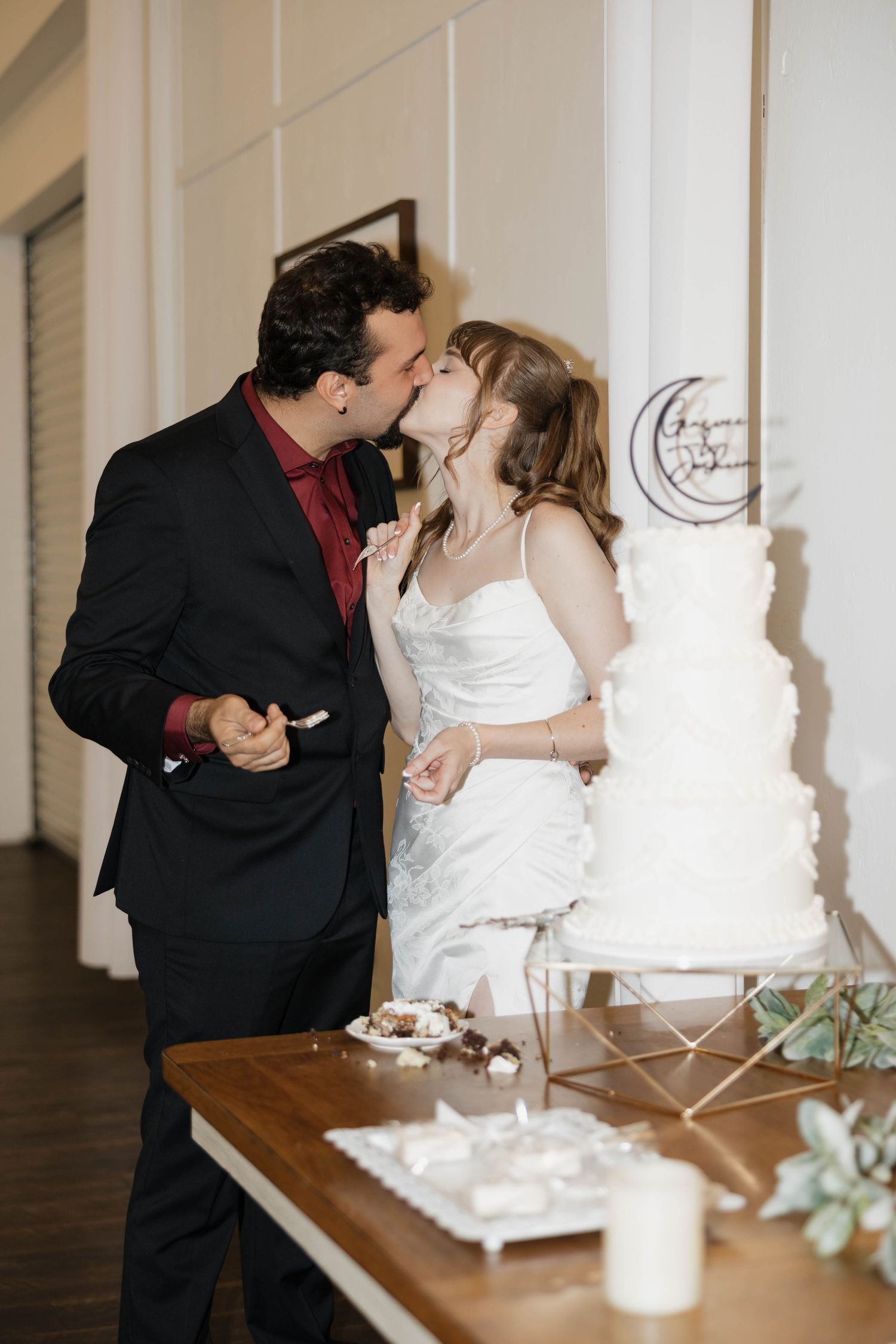 Bride and groom share a kiss while cutting their tiered white wedding cake at a reception dessert table.