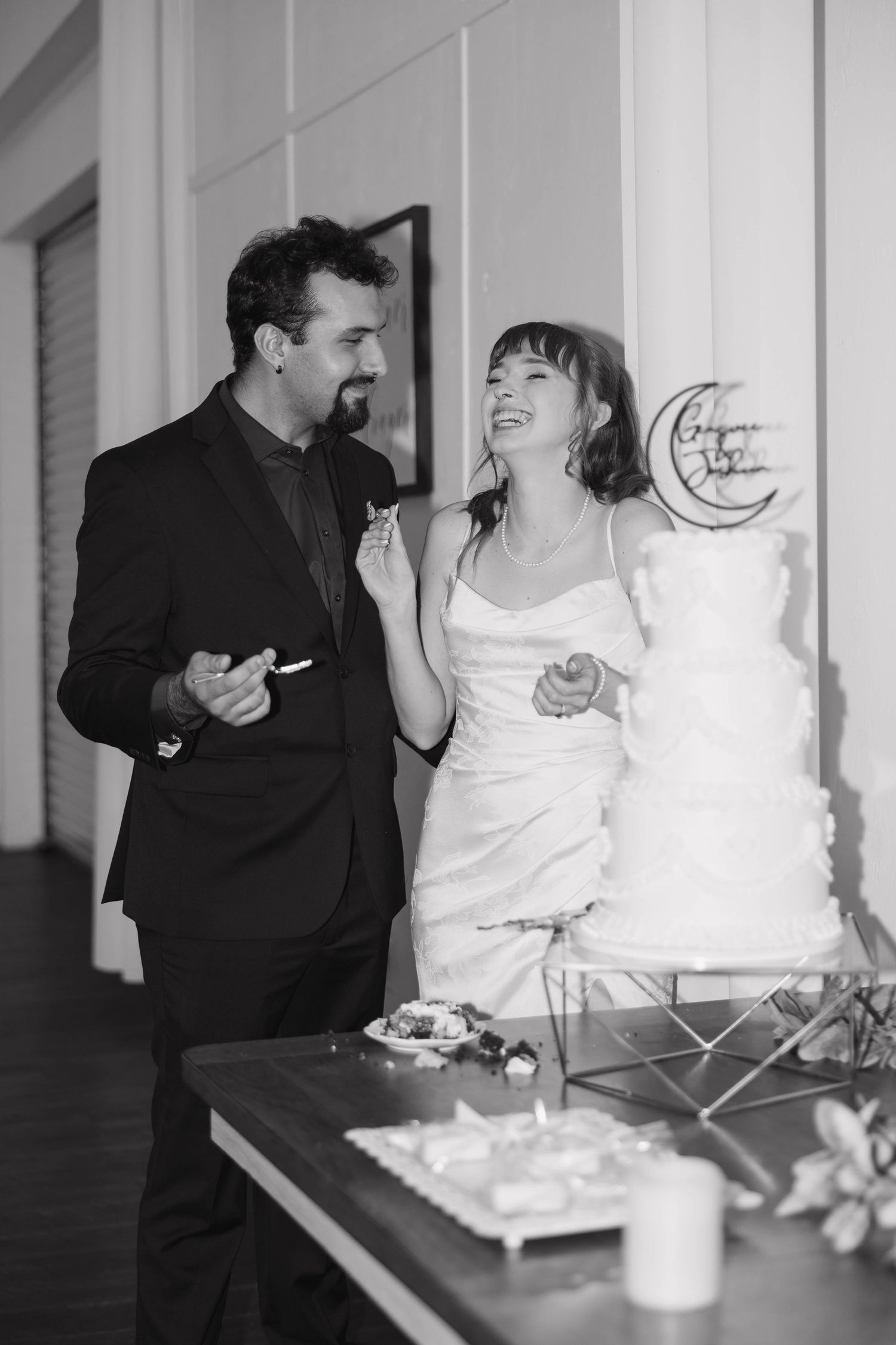 In a black and white photo, a laughing bride and groom share a bite of cake beside their tiered wedding cake with a moon topper.
