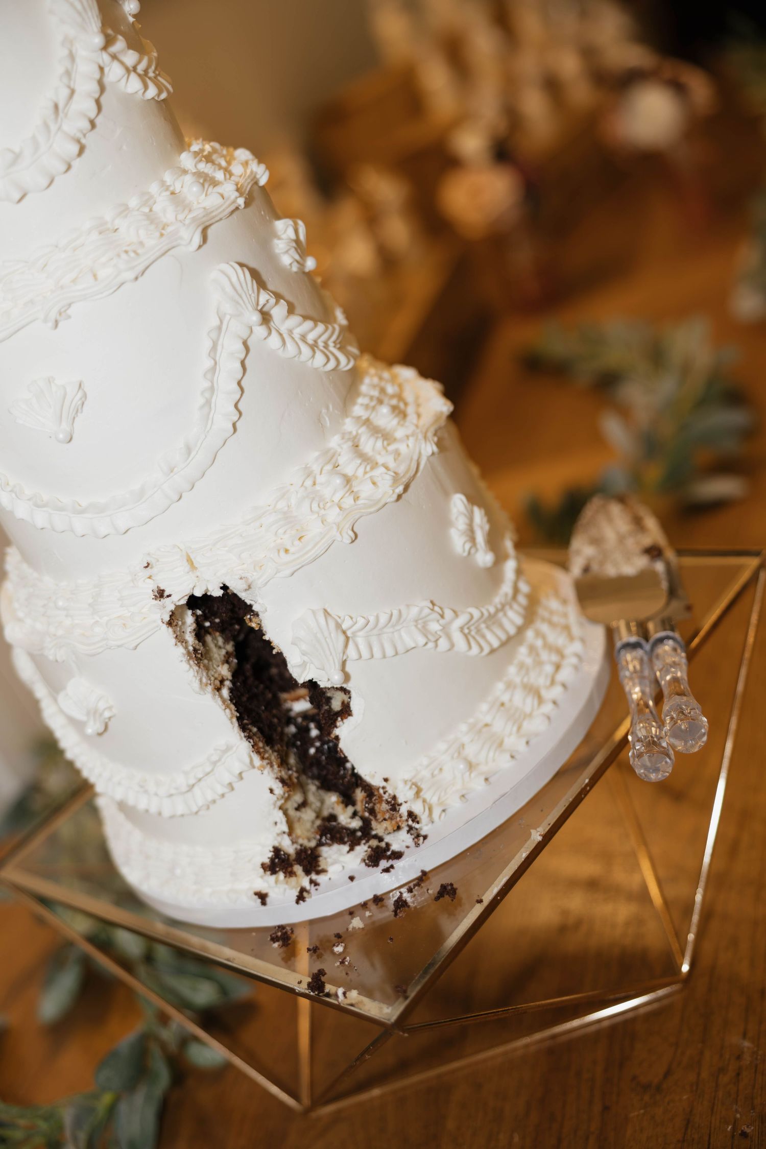 White tiered wedding cake with piped buttercream scrollwork and a slice cut out, revealing chocolate cake, on a geometric gold stand.