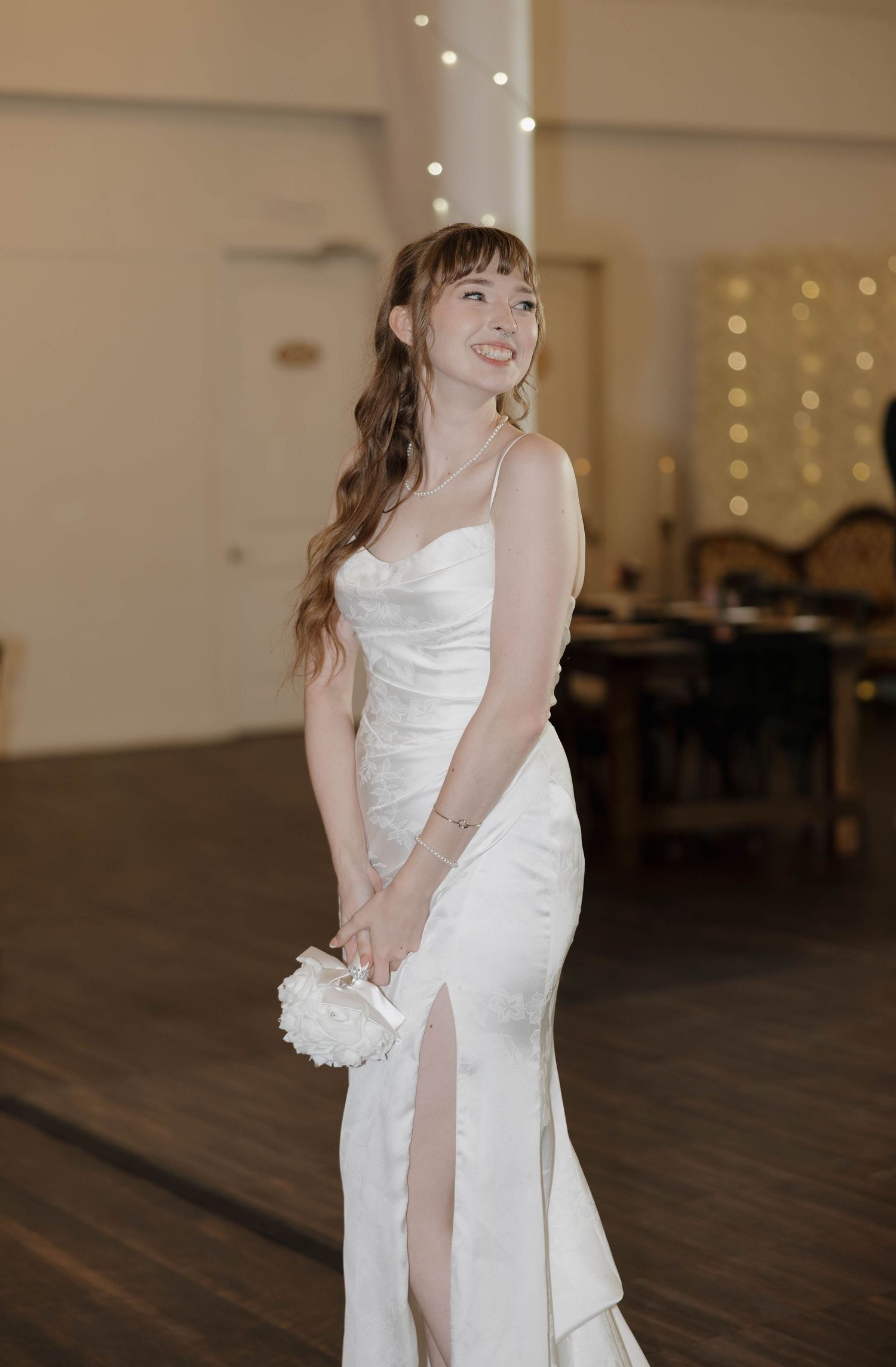 Smiling woman with long wavy hair and bangs wears a white satin slip gown with a high slit and holds a small white floral clutch indoors.