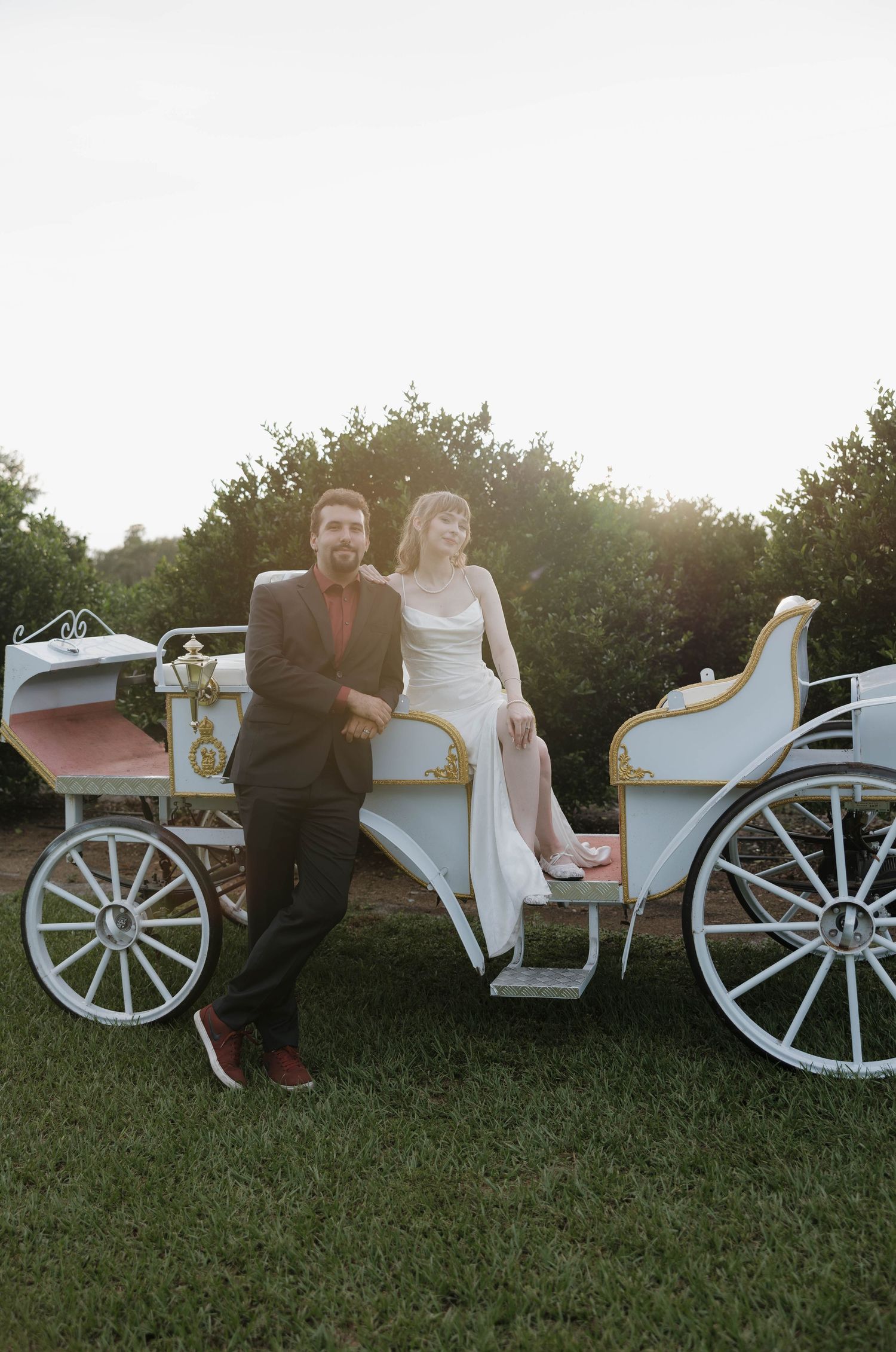 Bride in a white gown and groom in a dark suit pose beside a vintage white wedding carriage on a green lawn at sunset.