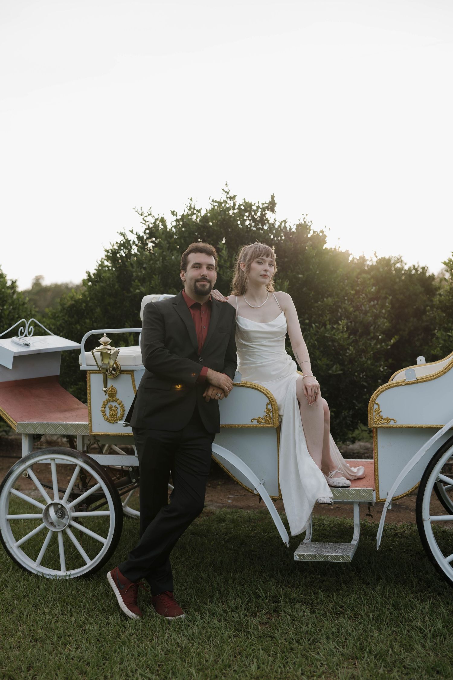 Bride in a satin white dress and groom in a dark suit pose together beside a white vintage horse-drawn carriage on a green lawn.