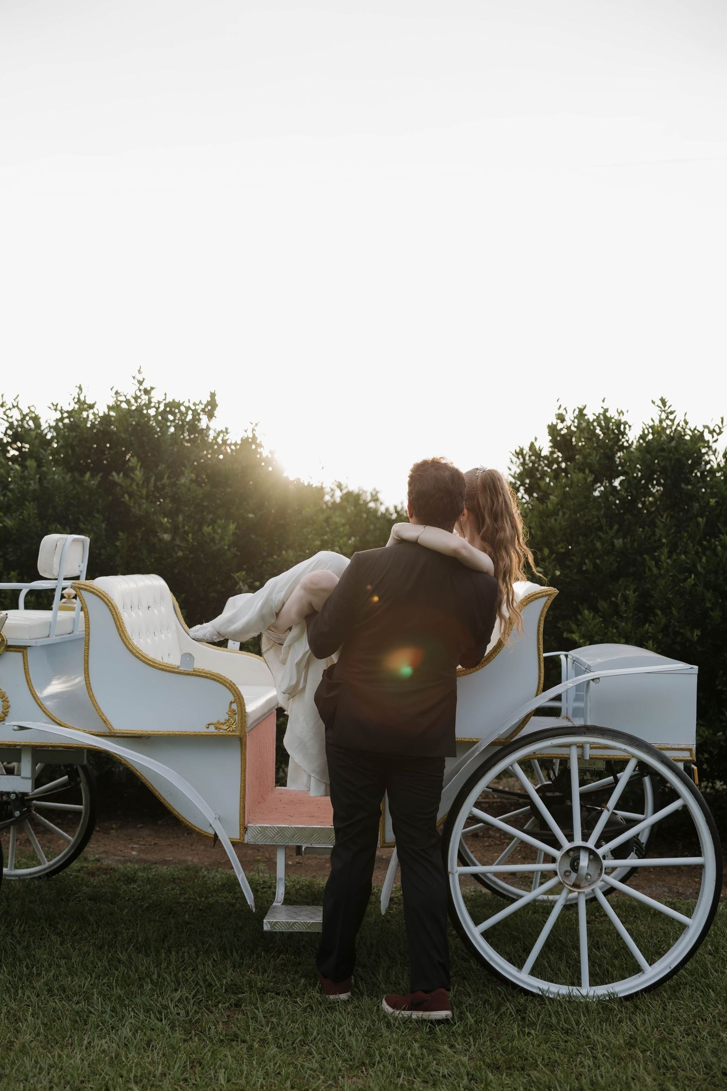 Groom holds his bride beside a white vintage horse carriage at sunset in a grassy outdoor setting.