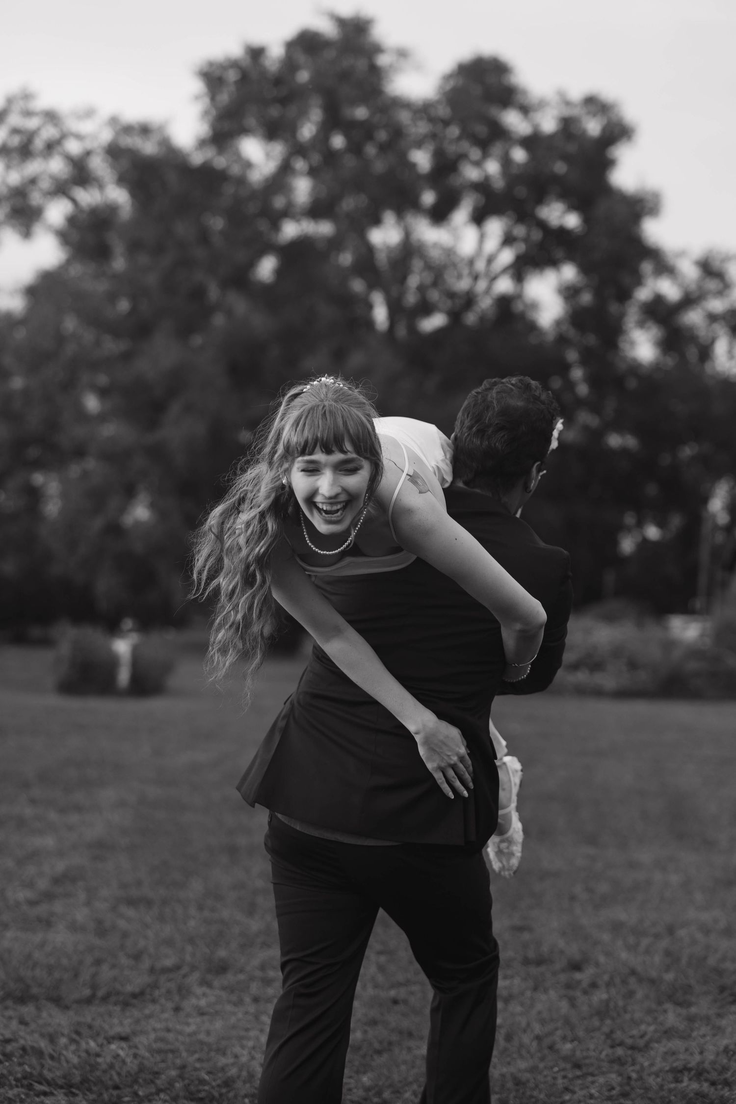 Laughing bride with long wavy hair and a tiara is carried over a groom's shoulder across a grassy park in a black and white wedding photo.