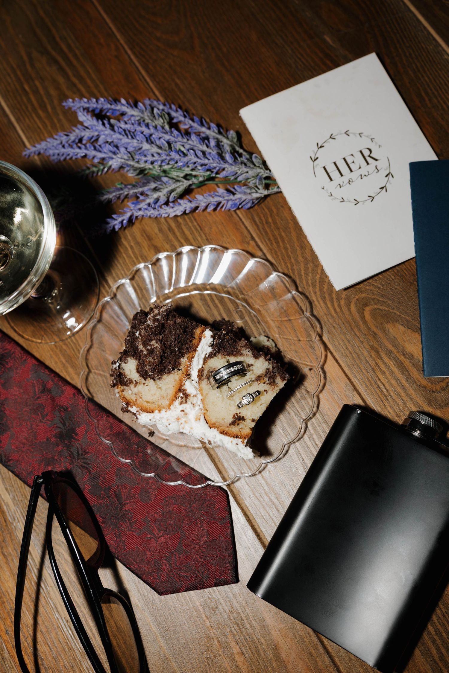 Overhead view of wedding cake slice with rings on a plate beside lavender, a burgundy tie, glasses, a flask and a vow booklet.