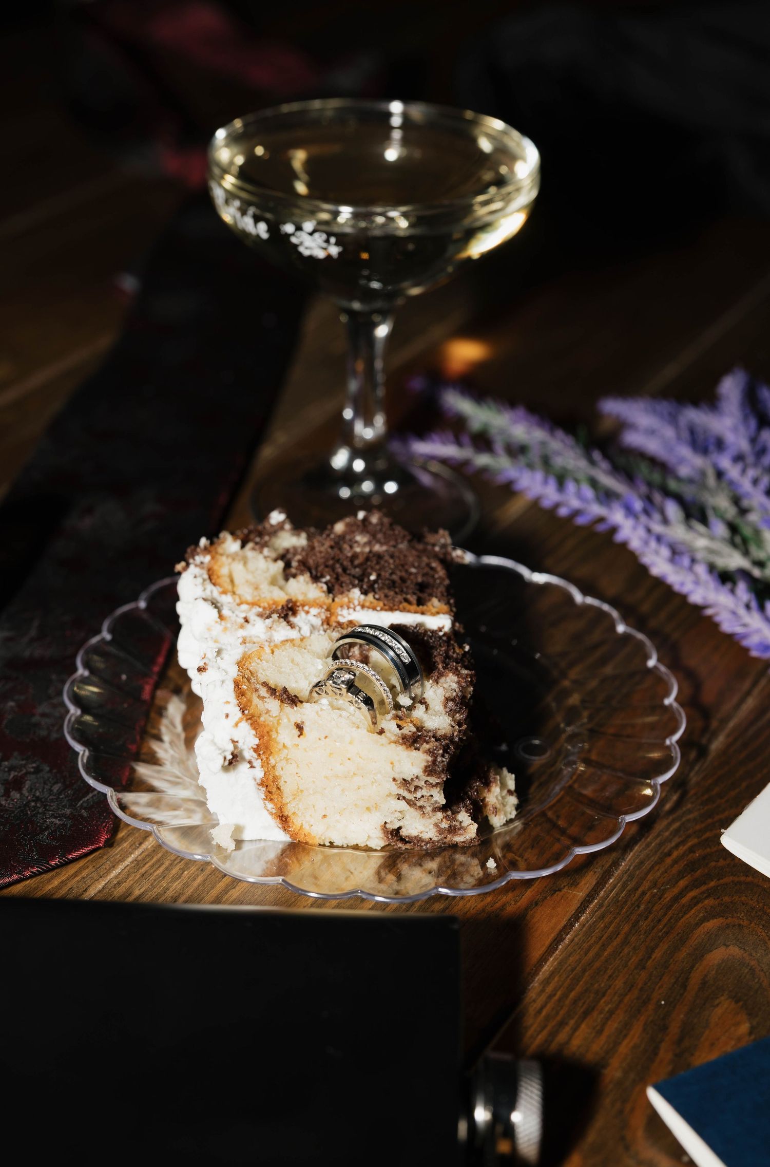 Slice of layered cake with white frosting and chocolate crumbs on a glass plate beside a champagne coupe and lavender on a wooden table.