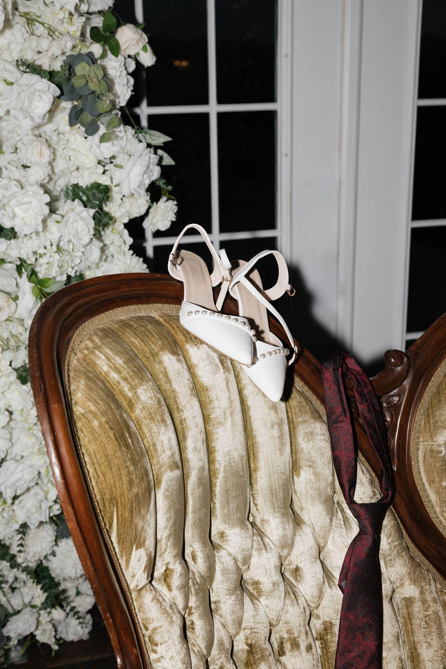White pearl-embellished wedding heels and a burgundy tie rest on a vintage tufted velvet chair beside white floral arrangements.
