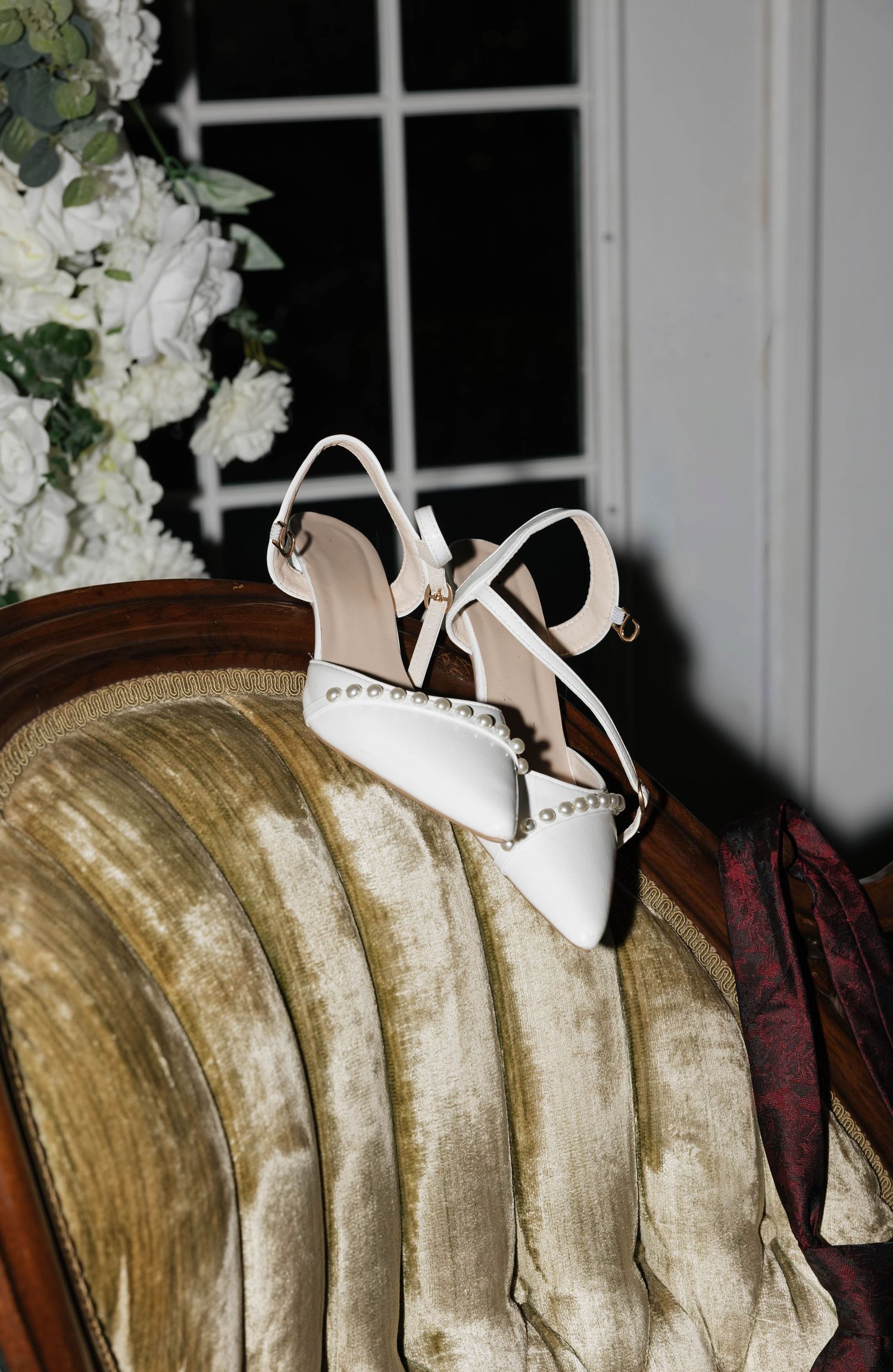 White pointed-toe bridal heels with pearl embellishments and ankle straps resting on a green velvet chair beside white floral arrangements.