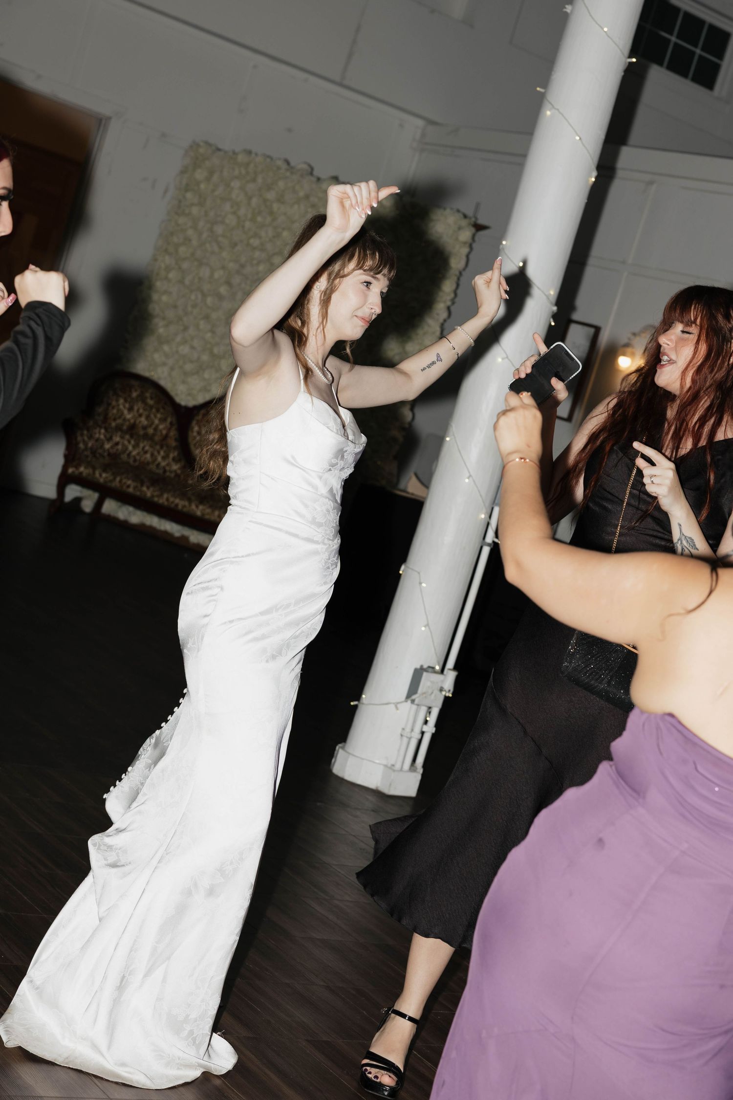 Woman in a white satin gown dances with raised arms at a wedding reception while guests in black and purple dresses dance nearby.