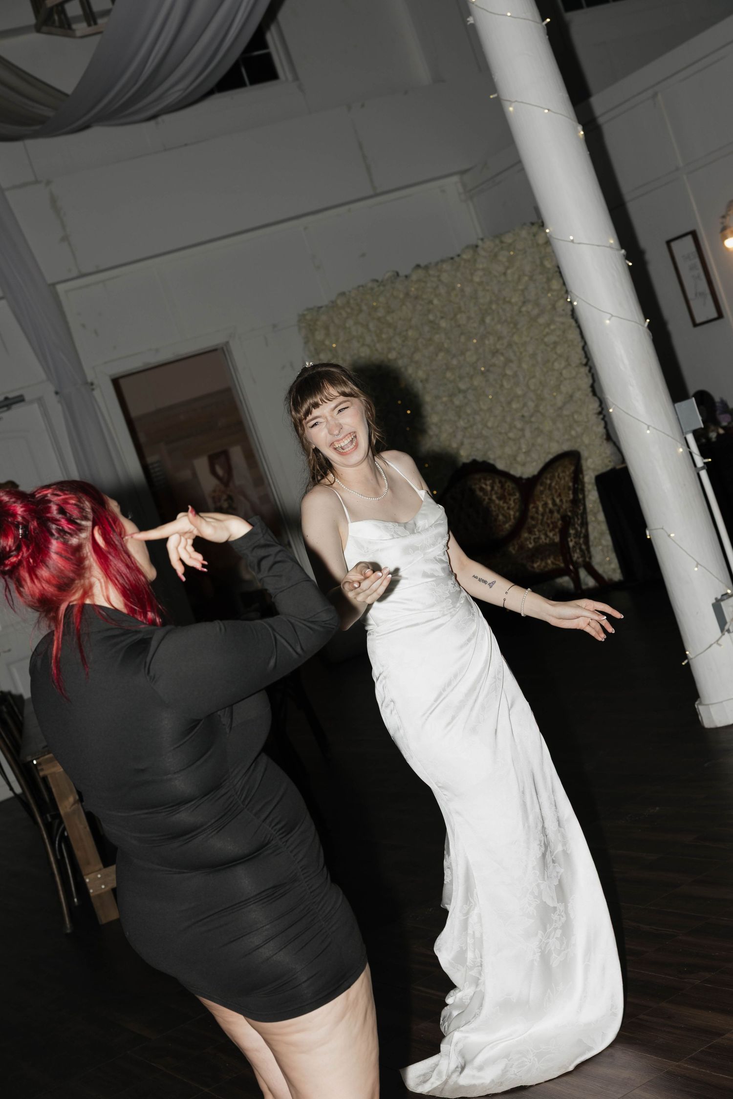 A bride in a white satin gown dances and laughs with a guest in a black dress at a wedding reception in a bright, airy venue.
