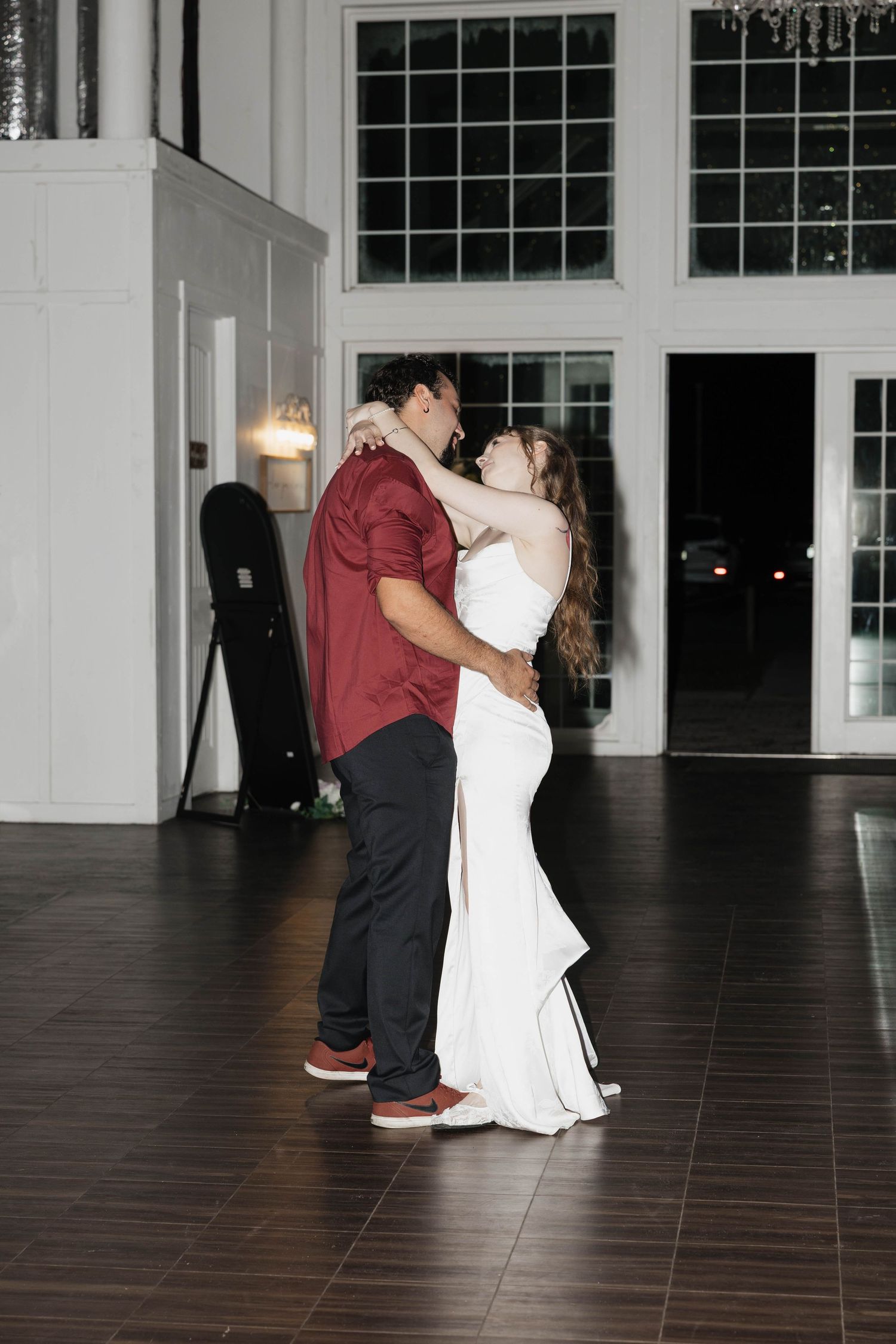 Bride in a white gown and groom in a red shirt share a slow dance on a dark wood floor at their wedding reception.