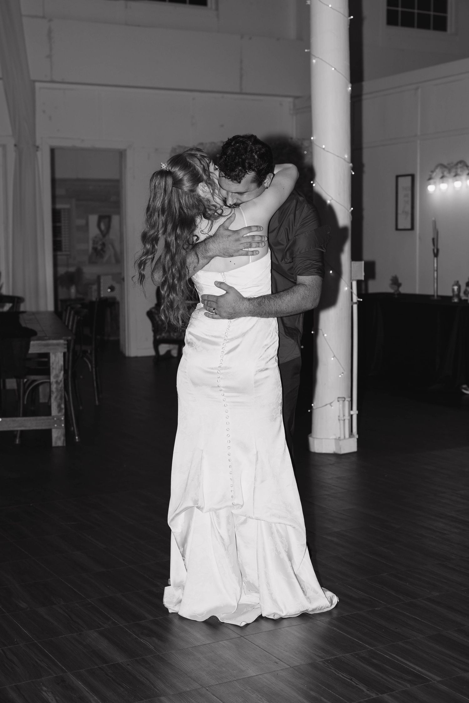 A black and white photo of a bride and groom sharing an intimate embrace on the dance floor of a rustic wedding reception venue.
