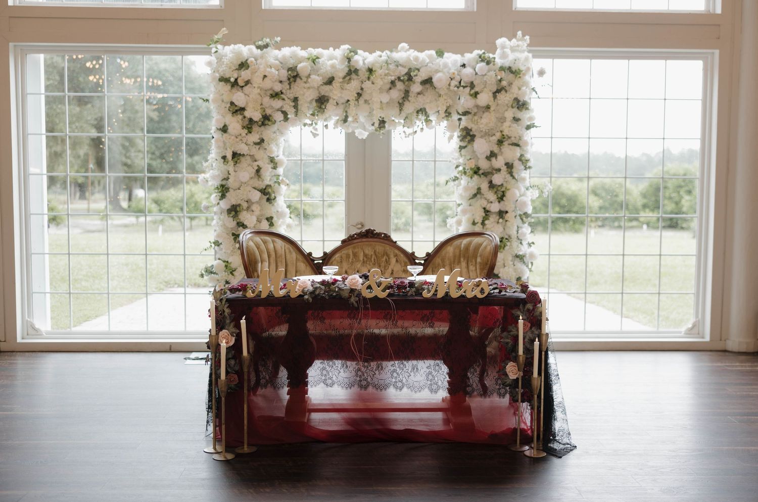 Elegant wedding sweetheart table with ornate gold chairs beneath a white floral arch, set before tall windows overlooking a green lawn.