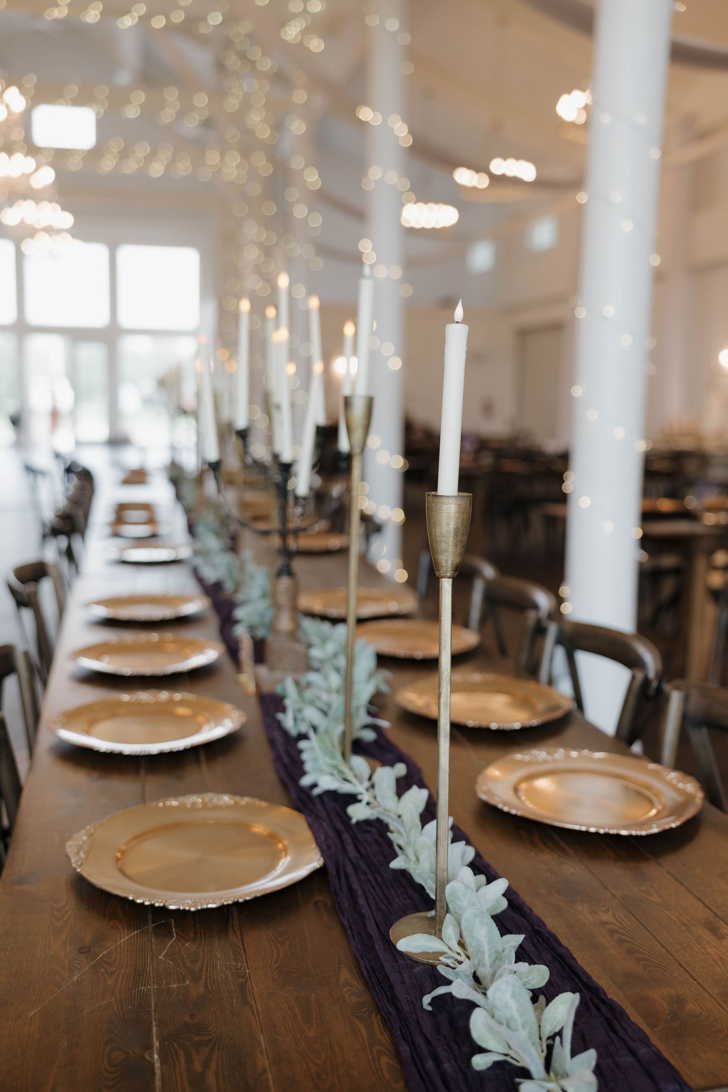 Long wooden reception table set with gold chargers, tall white taper candles, greenery, and a deep purple runner under string lights.